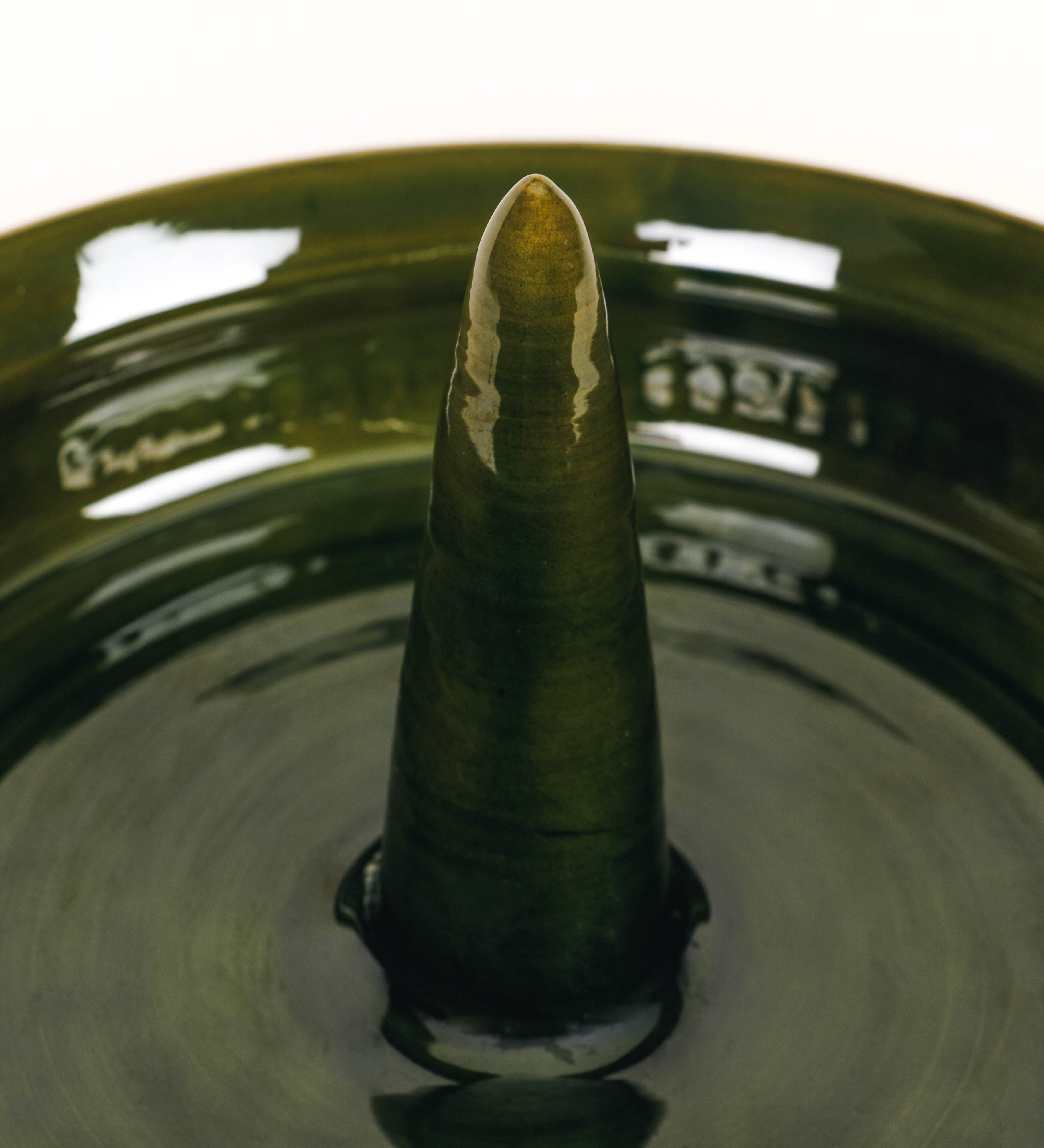 Assadeira para frangos com um design rústico e distinto. O assa-frangos é de barro, vidrado em tom verde vidro com brilho, conferindo um caráter vintage à peça. A forma do assa-frangos é circular, com uma borda superior mais estreita e um espeto cónico centrado no interior para colocar os frangos. A superfície externa da peça é decorada com um padrão de relevos losangulares, que envolvem o assa-frangos e acrescentam textura e interesse visual ao design.