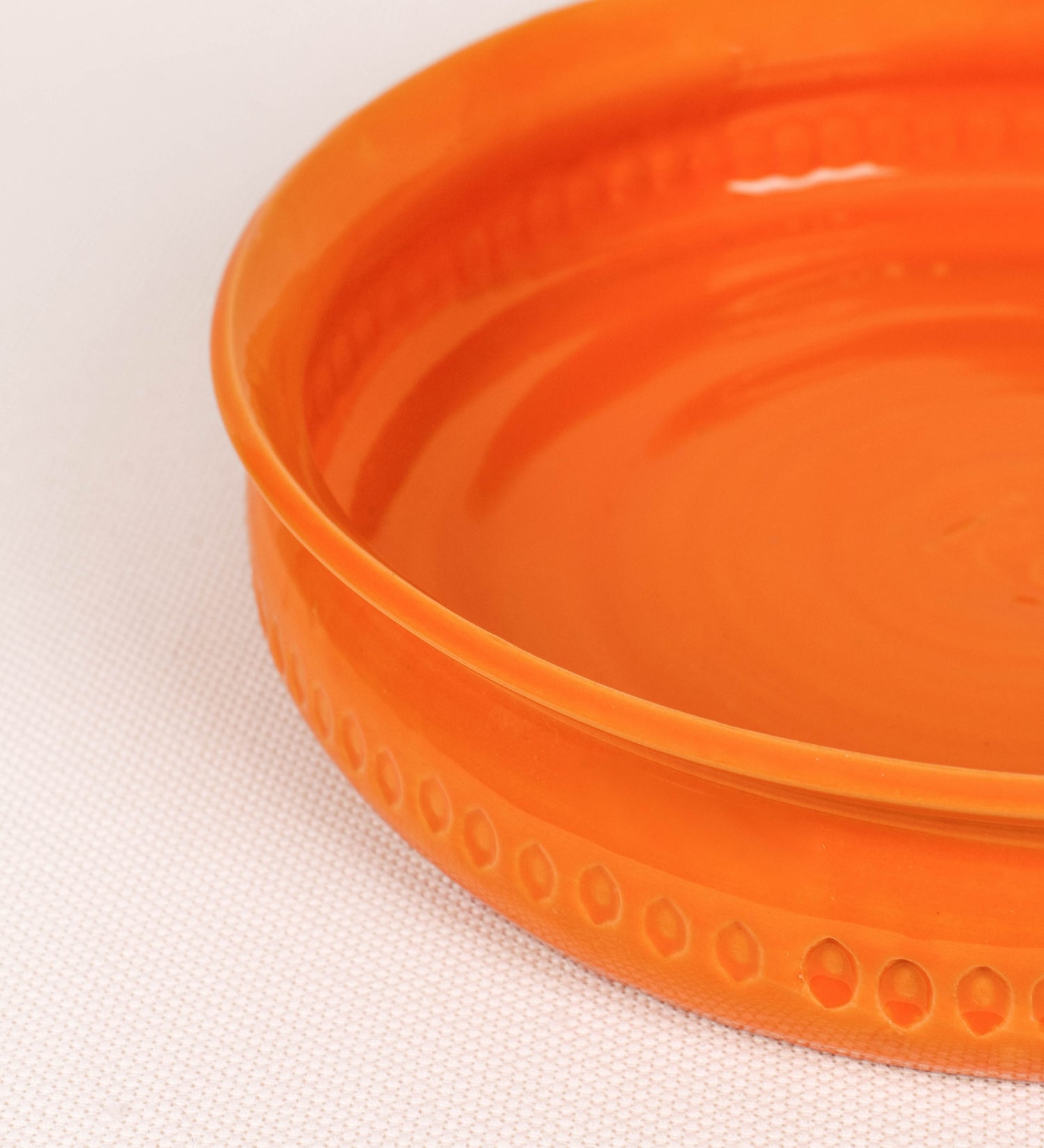 Assadeira com um design rústico e distinto. A assadeira é de barro, vidrado em tom laranja tangerina com brilho, conferindo um caráter chamativo à peça. A forma da assadeira é circular, com uma borda superior mais estreita. A superfície externa da peça é decorada com um padrão de relevos losangulares, que envolvem a assadeira e acrescentam textura e interesse visual ao design.