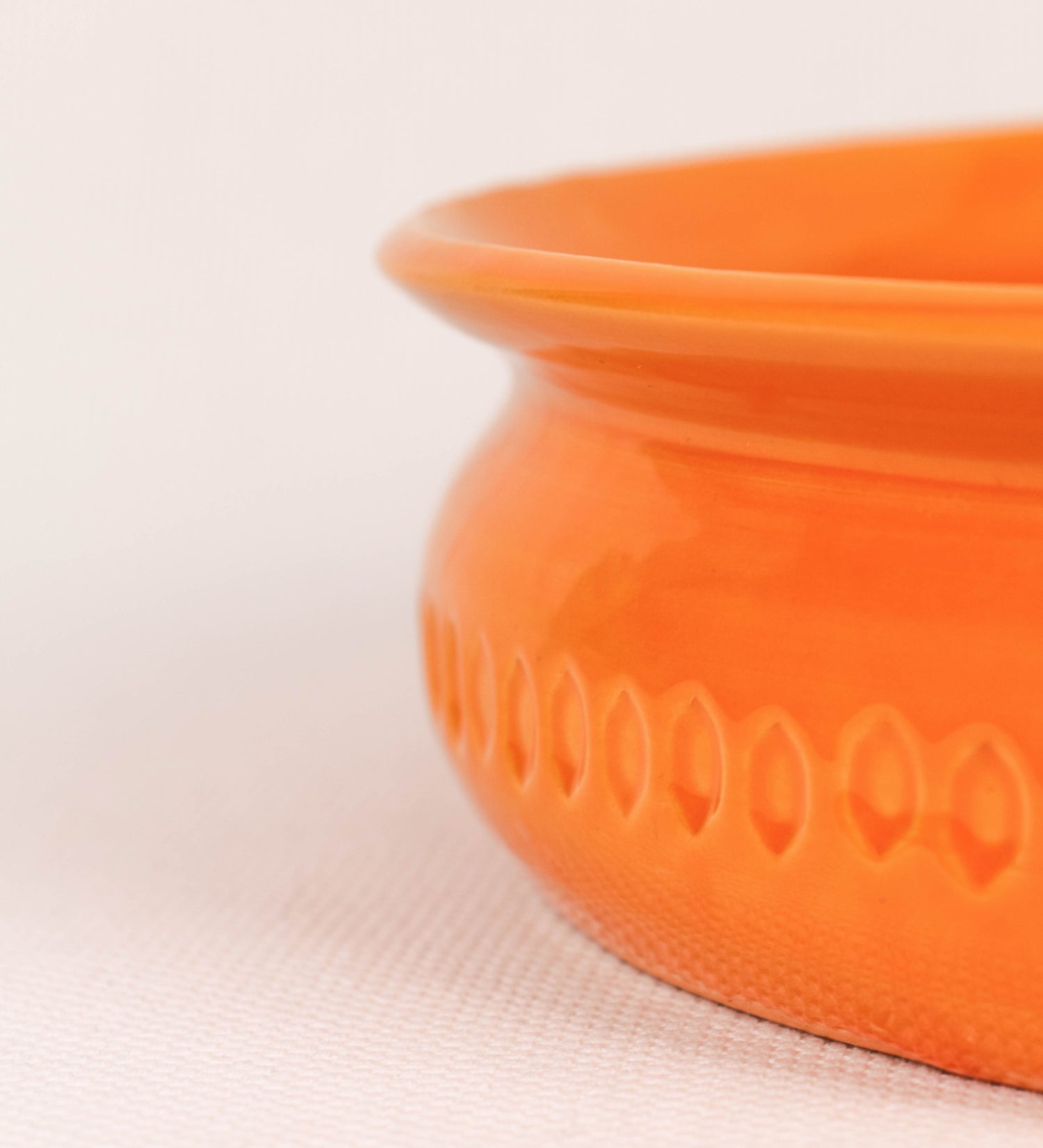 Assadeira com um design rústico e distinto. A assadeira é de barro, vidrado em tom laranja tangerina com brilho, conferindo um caráter chamativo à peça. A forma da assadeira é circular, com uma borda superior mais estreita. A superfície externa da peça é decorada com um padrão de relevos losangulares, que envolvem a assadeira e acrescentam textura e interesse visual ao design.
