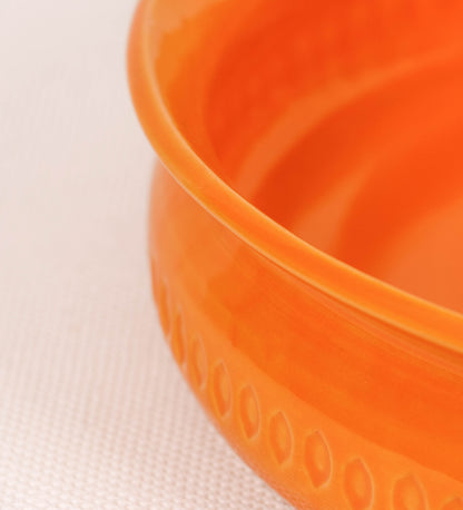 Assadeira com um design rústico e distinto. A assadeira é de barro, vidrado em tom laranja tangerina com brilho, conferindo um caráter chamativo à peça. A forma da assadeira é circular, com uma borda superior mais estreita. A superfície externa da peça é decorada com um padrão de relevos losangulares, que envolvem a assadeira e acrescentam textura e interesse visual ao design.