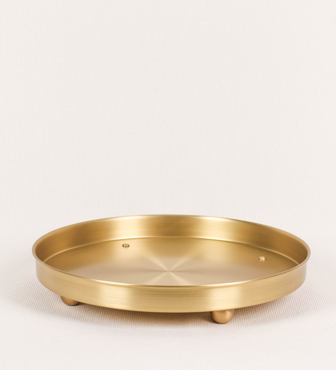 Bandeja com um design sofisticado e intemporal. A bandeja é de latão, com um tom dourado mate e acabamento escovado. Tem uma forma circular, de superfície plana e com uma borda envolvente de altura média e grossura fina. Na sua base tem três pequenas esferas, em latão também, dispostas em formato triangular, que sustentam o prato circular no topo. 