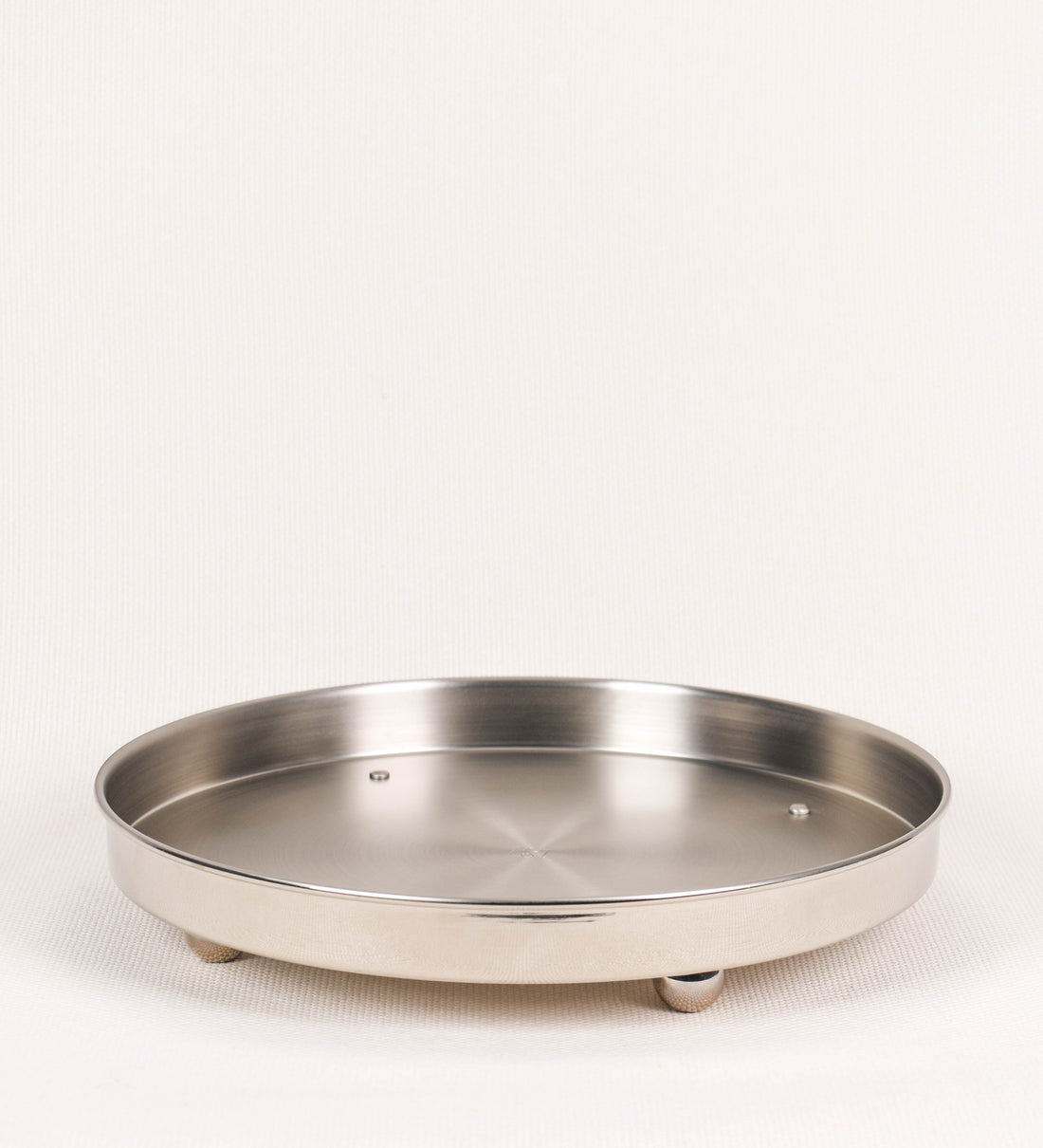 Bandeja com um design sofisticado e intemporal. A bandeja é de latão, tendo na superfície exterior um acabamento em tom prateado e com efeito de espelho, e um acabamento em tom prateado com efeito mate na superfície interior. Tem uma forma circular, de superfície plana e com uma borda envolvente de altura média e grossura fina. Na sua base tem três pequenas esferas, em níquel polido também, dispostas em formato triangular, que sustentam o prato circular no topo. 