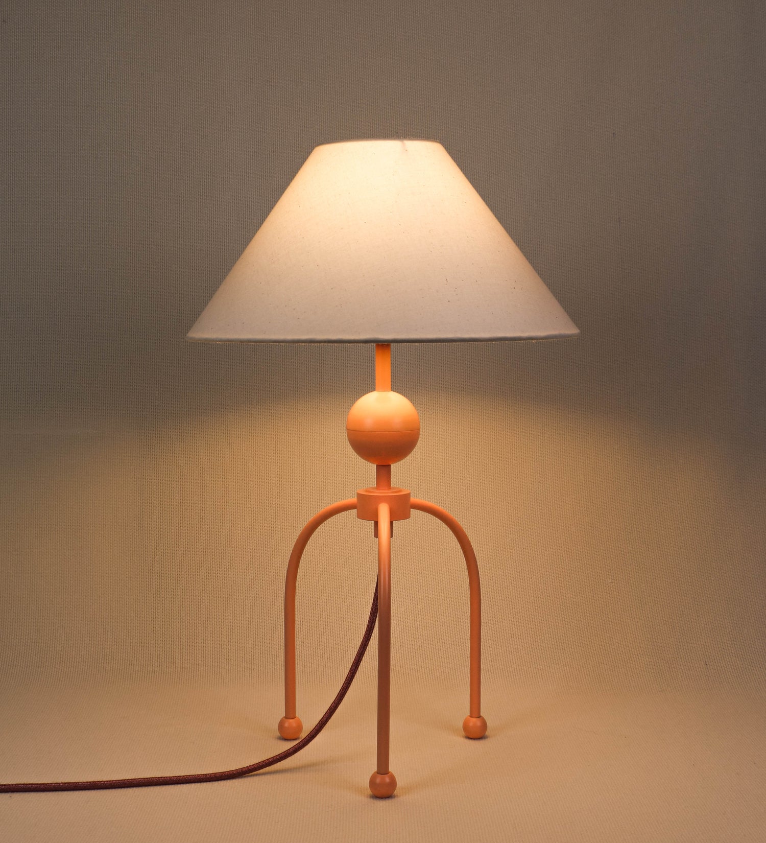 Candeeiro de mesa, de tom rosa salmão, com um design moderno e minimalista. O candeeiro tem o corpo com três pés curvados, terminando em pequenas esferas. A estrutura dos pés é interligada por uma peça central esférica. O cabo é revestido têxtil de tom rosa velho, com interruptor e ficha transparentes. O abatjour é de forma cónica, em algodão, de tom bege claro com pequenas pintas escuras, semelhante à baunilha.