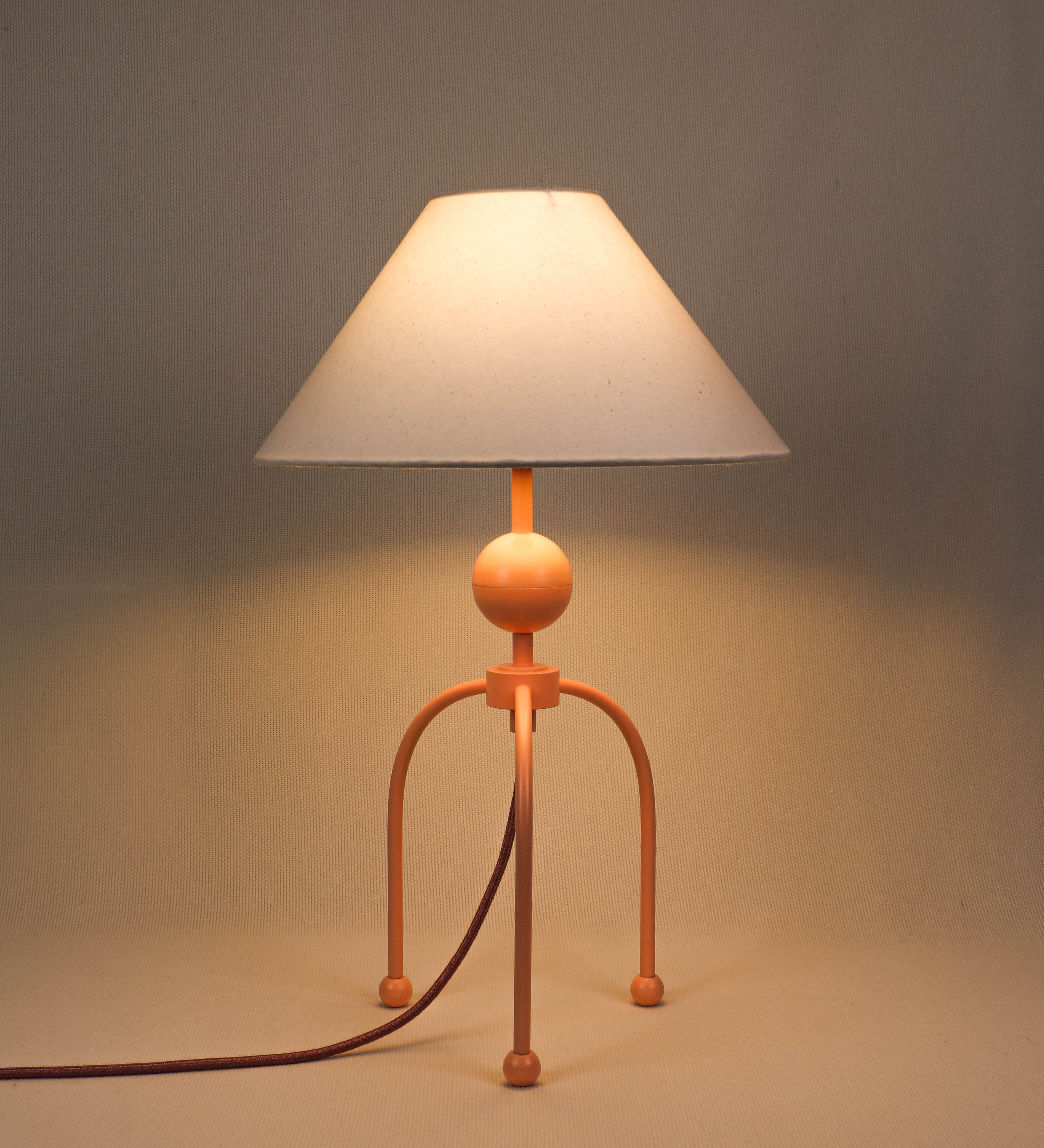 Candeeiro de mesa, de tom rosa salmão, com um design moderno e minimalista. O candeeiro tem o corpo com três pés curvados, terminando em pequenas esferas. A estrutura dos pés é interligada por uma peça central esférica. O cabo é revestido têxtil de tom rosa velho, com interruptor e ficha transparentes. O abatjour é de forma cónica, em algodão, de tom bege claro com pequenas pintas escuras, semelhante à baunilha.