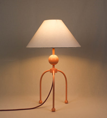Candeeiro de mesa, de tom rosa salmão, com um design moderno e minimalista. O candeeiro tem o corpo com três pés curvados, terminando em pequenas esferas. A estrutura dos pés é interligada por uma peça central esférica. O cabo é revestido têxtil de tom rosa velho, com interruptor e ficha transparentes. O abatjour é de forma cónica, em algodão, de tom bege claro com pequenas pintas escuras, semelhante à baunilha.