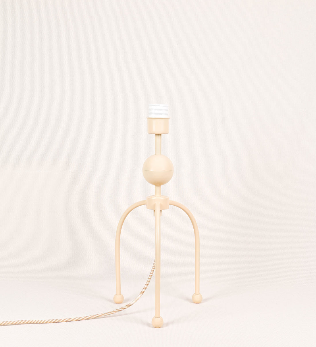 Candeeiro de mesa, de tom bege claro, com um design moderno e minimalista. O candeeiro tem o corpo com três pés curvados, terminando em pequenas esferas. A estrutura dos pés é interligada por uma peça central esférica. O cabo é revestido têxtil com um padrão de linhas douradas e brancas entrelaçadas, com interruptor e ficha transparentes.