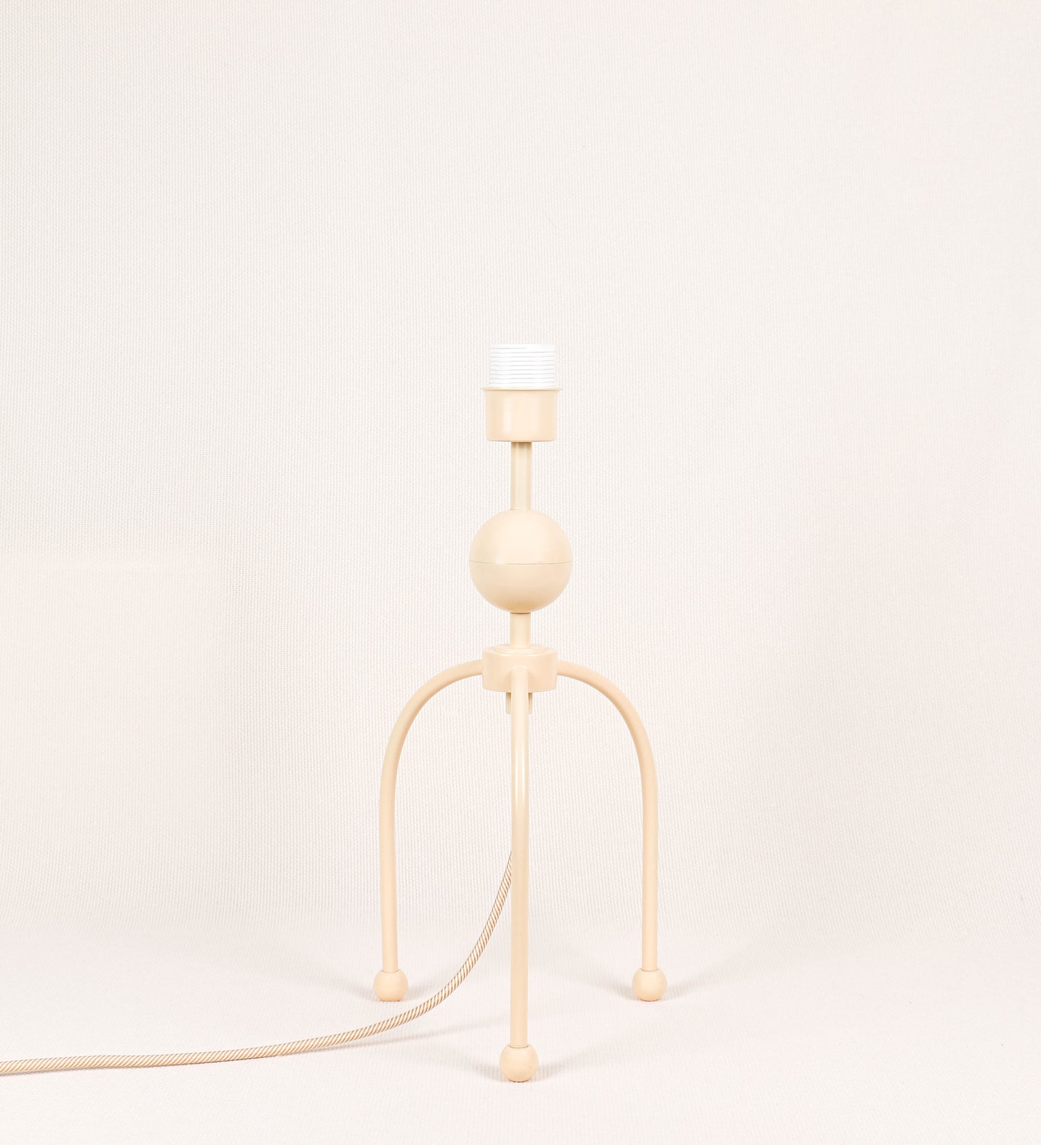 Candeeiro de mesa, de tom bege claro, com um design moderno e minimalista. O candeeiro tem o corpo com três pés curvados, terminando em pequenas esferas. A estrutura dos pés é interligada por uma peça central esférica. O cabo é revestido têxtil com um padrão de linhas douradas e brancas entrelaçadas, com interruptor e ficha transparentes.