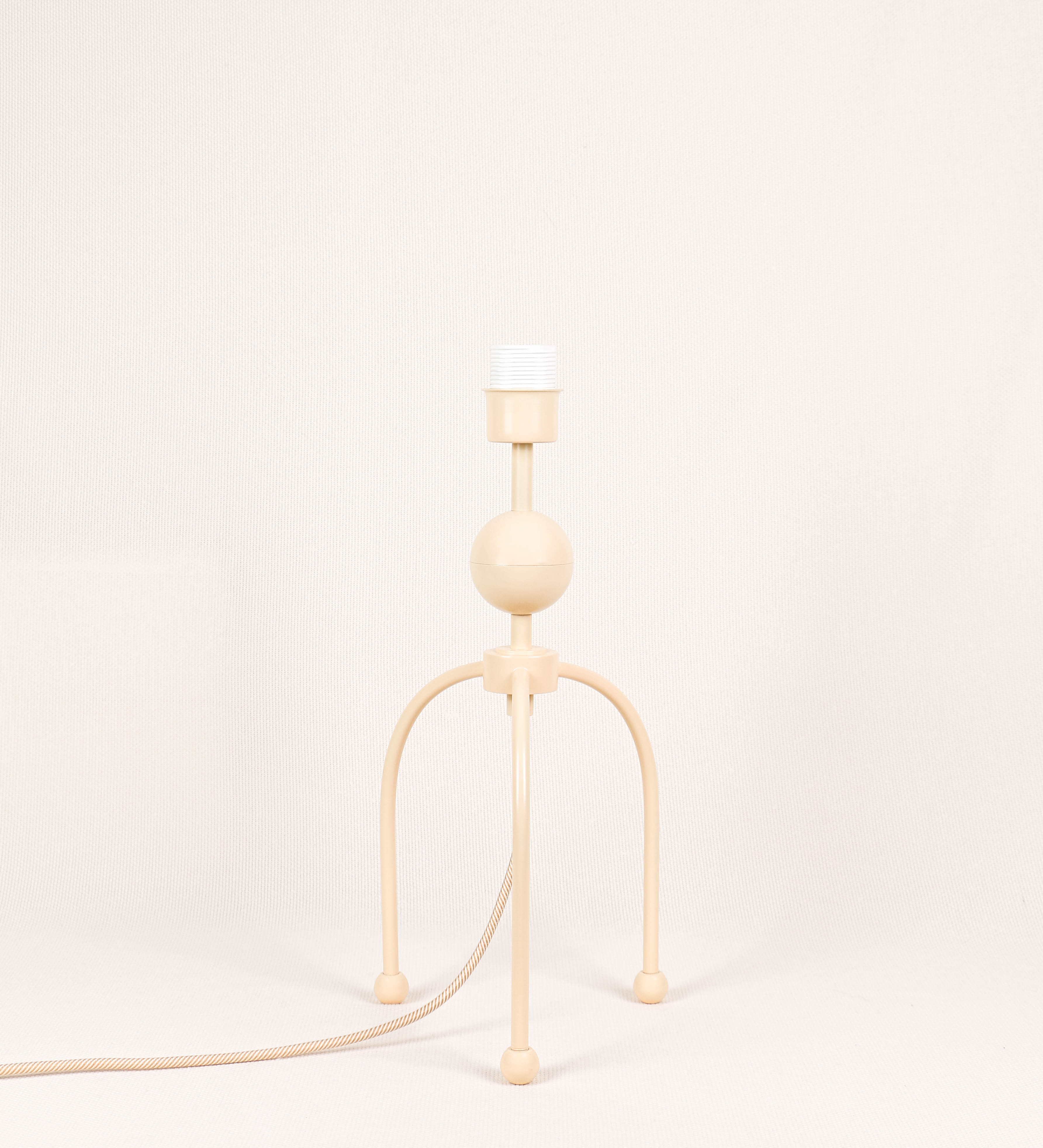 Candeeiro de mesa, de tom bege claro, com um design moderno e minimalista. O candeeiro tem o corpo com três pés curvados, terminando em pequenas esferas. A estrutura dos pés é interligada por uma peça central esférica. O cabo é revestido têxtil com um padrão de linhas douradas e brancas entrelaçadas, com interruptor e ficha transparentes.