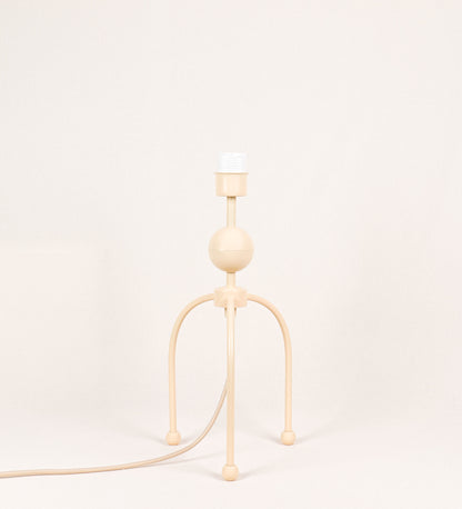 Candeeiro de mesa, de tom bege claro, com um design moderno e minimalista. O candeeiro tem o corpo com três pés curvados, terminando em pequenas esferas. A estrutura dos pés é interligada por uma peça central esférica. O cabo é revestido têxtil com um padrão de linhas douradas e brancas entrelaçadas, com interruptor e ficha transparentes.