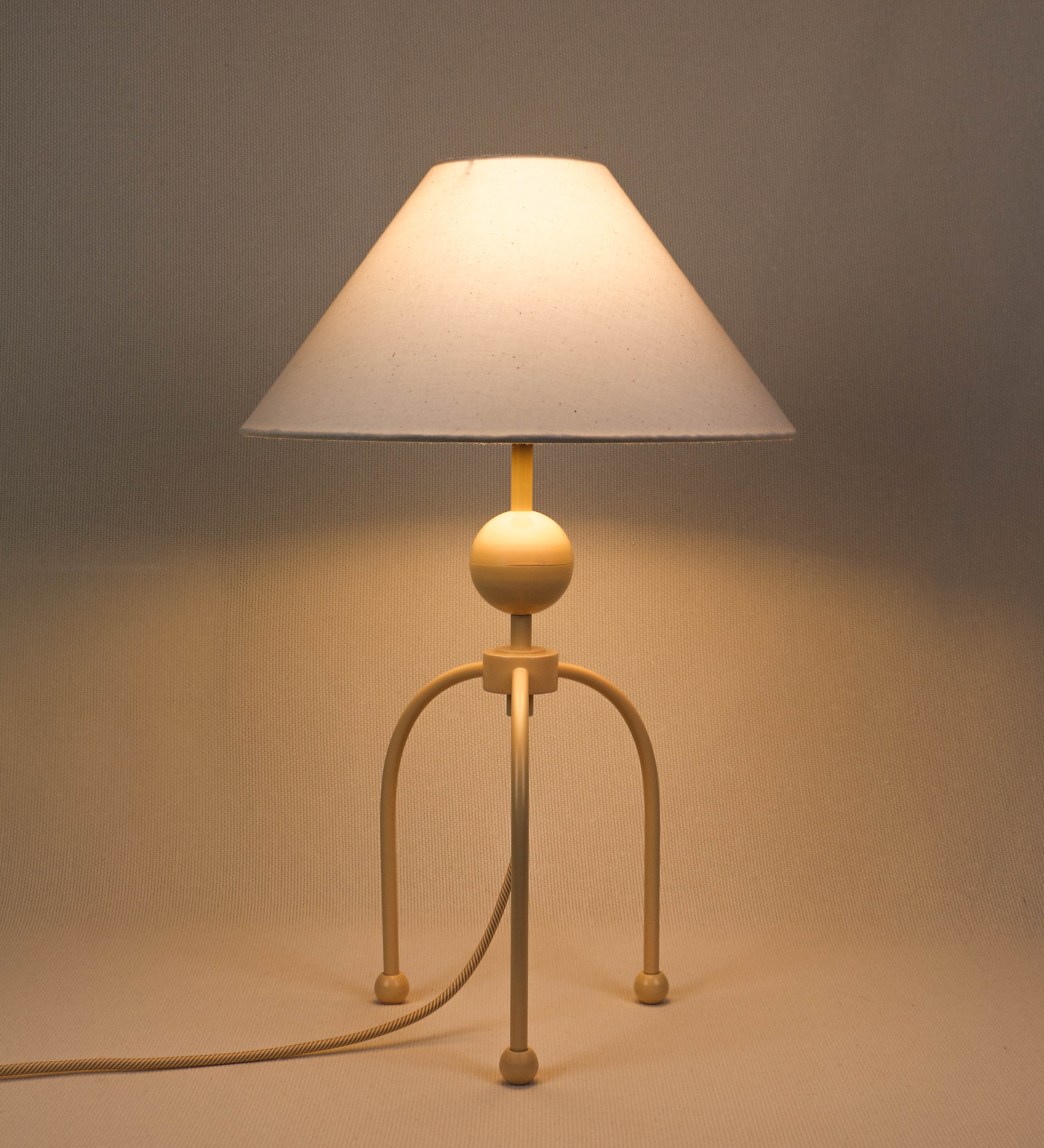 Candeeiro de mesa, de tom bege claro, com um design moderno e minimalista. O candeeiro tem o corpo com três pés curvados, terminando em pequenas esferas. A estrutura dos pés é interligada por uma peça central esférica. O cabo é revestido têxtil com um padrão de linhas douradas e brancas entrelaçadas, com interruptor e ficha transparentes. O abatjour é de forma cónica, em algodão, de tom bege claro com pequenas pintas escuras, semelhante à baunilha.