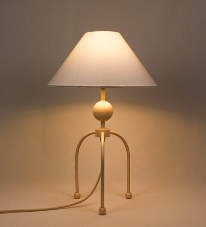 Candeeiro de mesa, de tom bege claro, com um design moderno e minimalista. O candeeiro tem o corpo com três pés curvados, terminando em pequenas esferas. A estrutura dos pés é interligada por uma peça central esférica. O cabo é revestido têxtil com um padrão de linhas douradas e brancas entrelaçadas, com interruptor e ficha transparentes. O abatjour é de forma cónica, em algodão, de tom bege claro com pequenas pintas escuras, semelhante à baunilha.