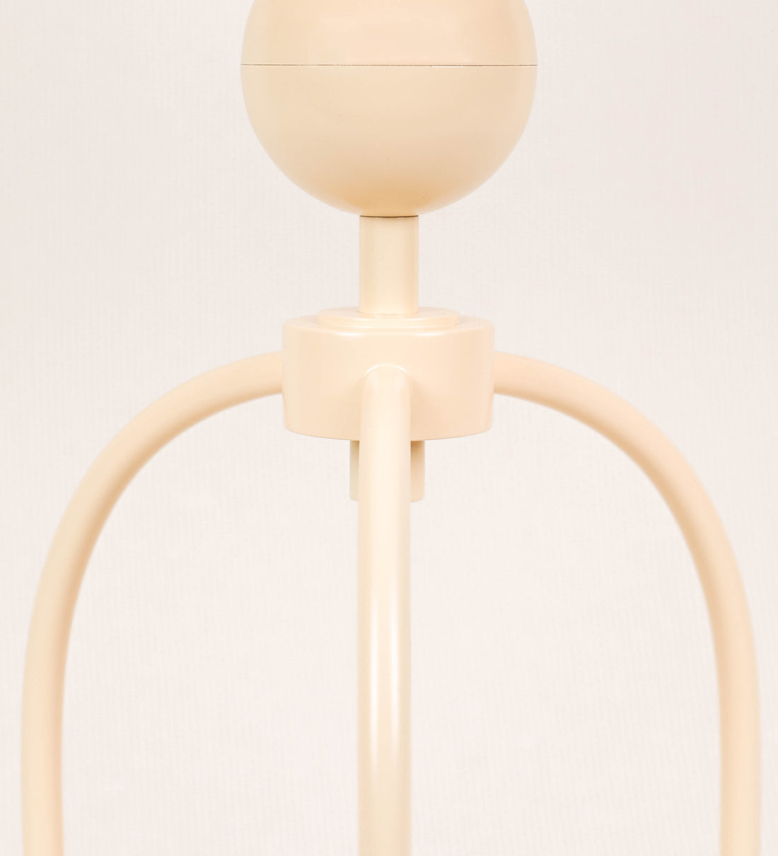 Candeeiro de mesa, de tom bege claro, com um design moderno e minimalista. O candeeiro tem o corpo com três pés curvados, terminando em pequenas esferas. A estrutura dos pés é interligada por uma peça central esférica. O cabo é revestido têxtil com um padrão de linhas douradas e brancas entrelaçadas, com interruptor e ficha transparentes.