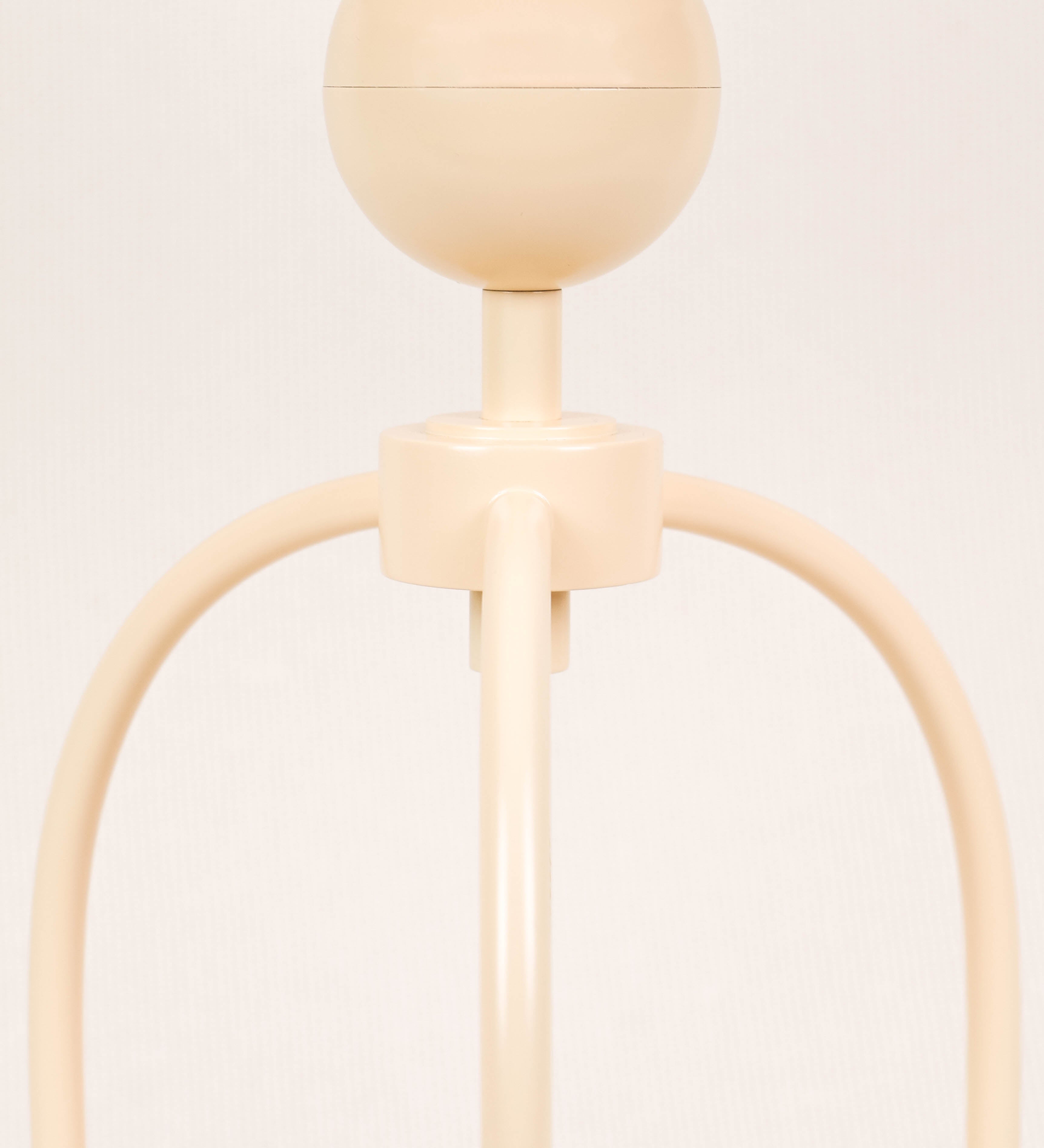 Candeeiro de mesa, de tom bege claro, com um design moderno e minimalista. O candeeiro tem o corpo com três pés curvados, terminando em pequenas esferas. A estrutura dos pés é interligada por uma peça central esférica. O cabo é revestido têxtil com um padrão de linhas douradas e brancas entrelaçadas, com interruptor e ficha transparentes.
