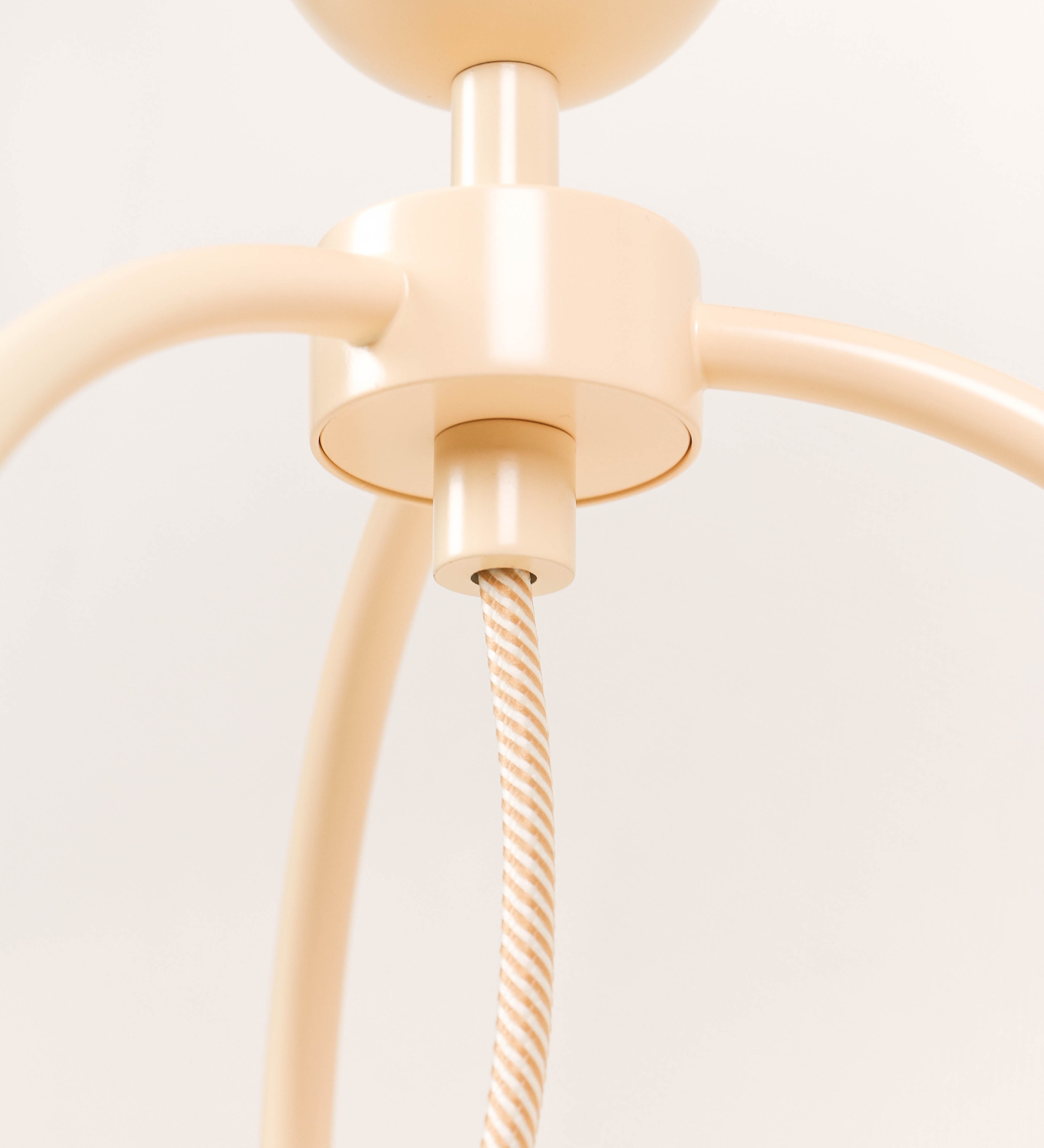 Candeeiro de mesa, de tom bege claro, com um design moderno e minimalista. O candeeiro tem o corpo com três pés curvados, terminando em pequenas esferas. A estrutura dos pés é interligada por uma peça central esférica. O cabo é revestido têxtil com um padrão de linhas douradas e brancas entrelaçadas, com interruptor e ficha transparentes.