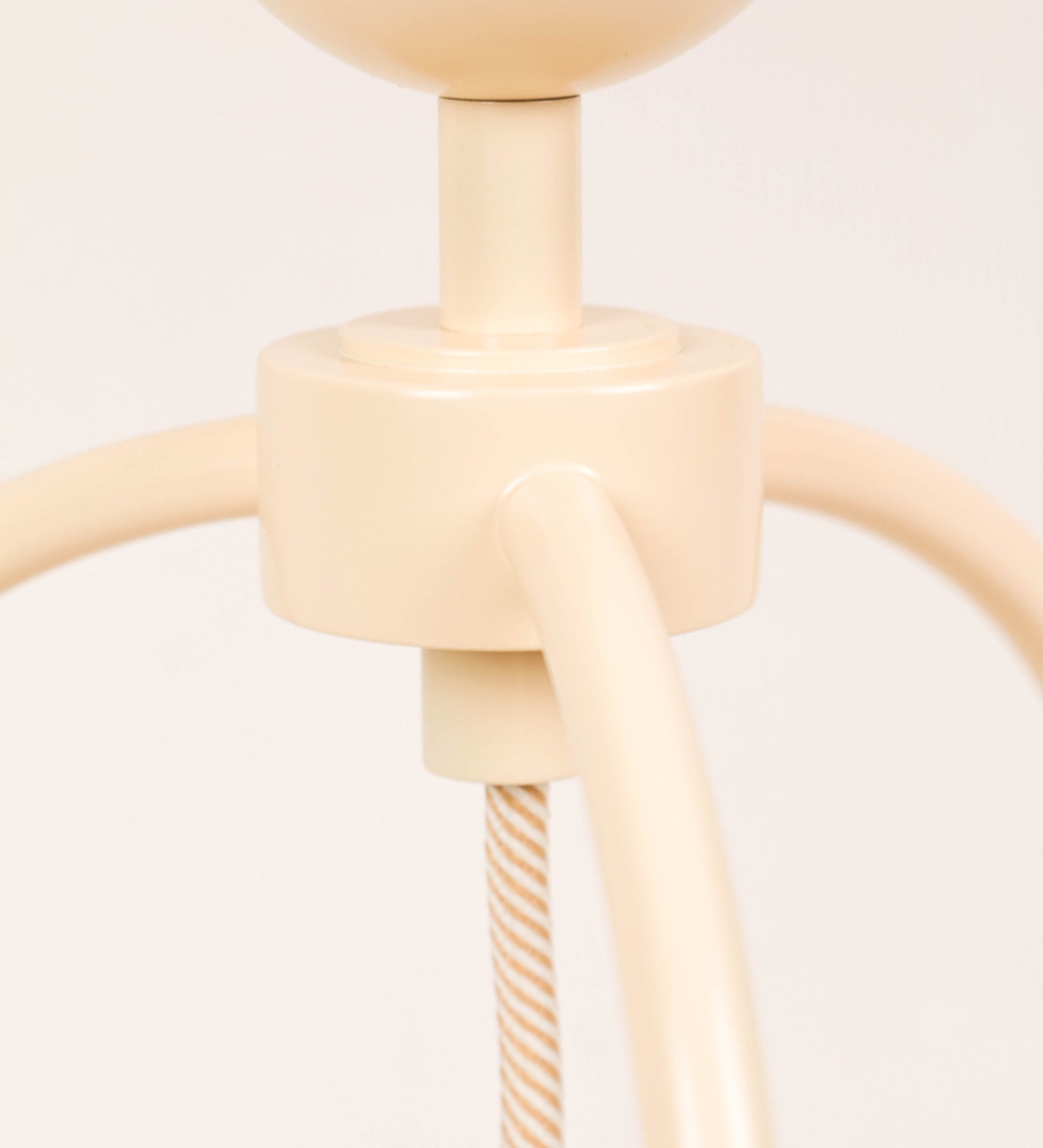 Candeeiro de mesa, de tom bege claro, com um design moderno e minimalista. O candeeiro tem o corpo com três pés curvados, terminando em pequenas esferas. A estrutura dos pés é interligada por uma peça central esférica. O cabo é revestido têxtil com um padrão de linhas douradas e brancas entrelaçadas, com interruptor e ficha transparentes.