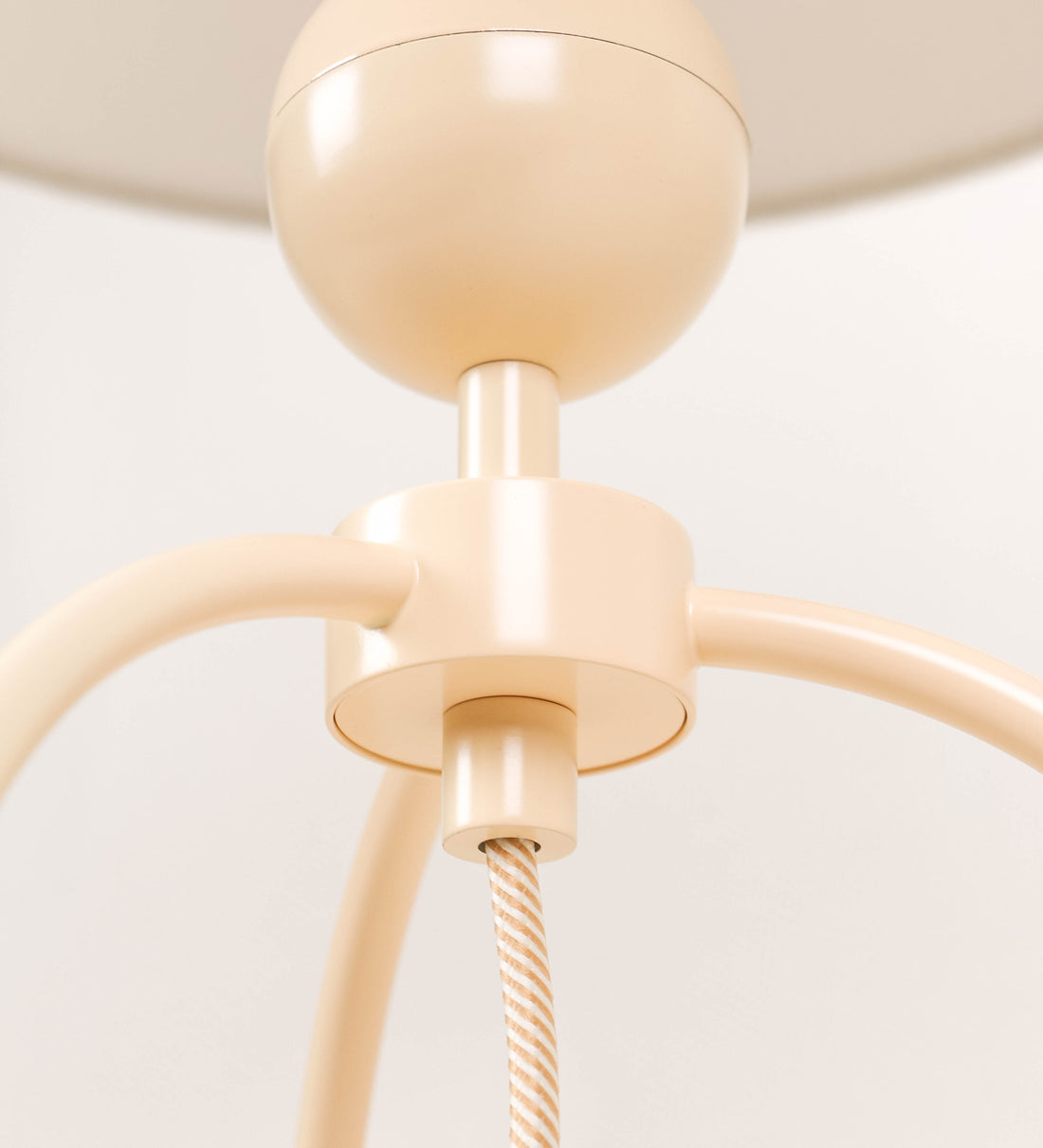 Candeeiro de mesa, de tom bege claro, com um design moderno e minimalista. O candeeiro tem o corpo com três pés curvados, terminando em pequenas esferas. A estrutura dos pés é interligada por uma peça central esférica. O cabo é revestido têxtil com um padrão de linhas douradas e brancas entrelaçadas, com interruptor e ficha transparentes.