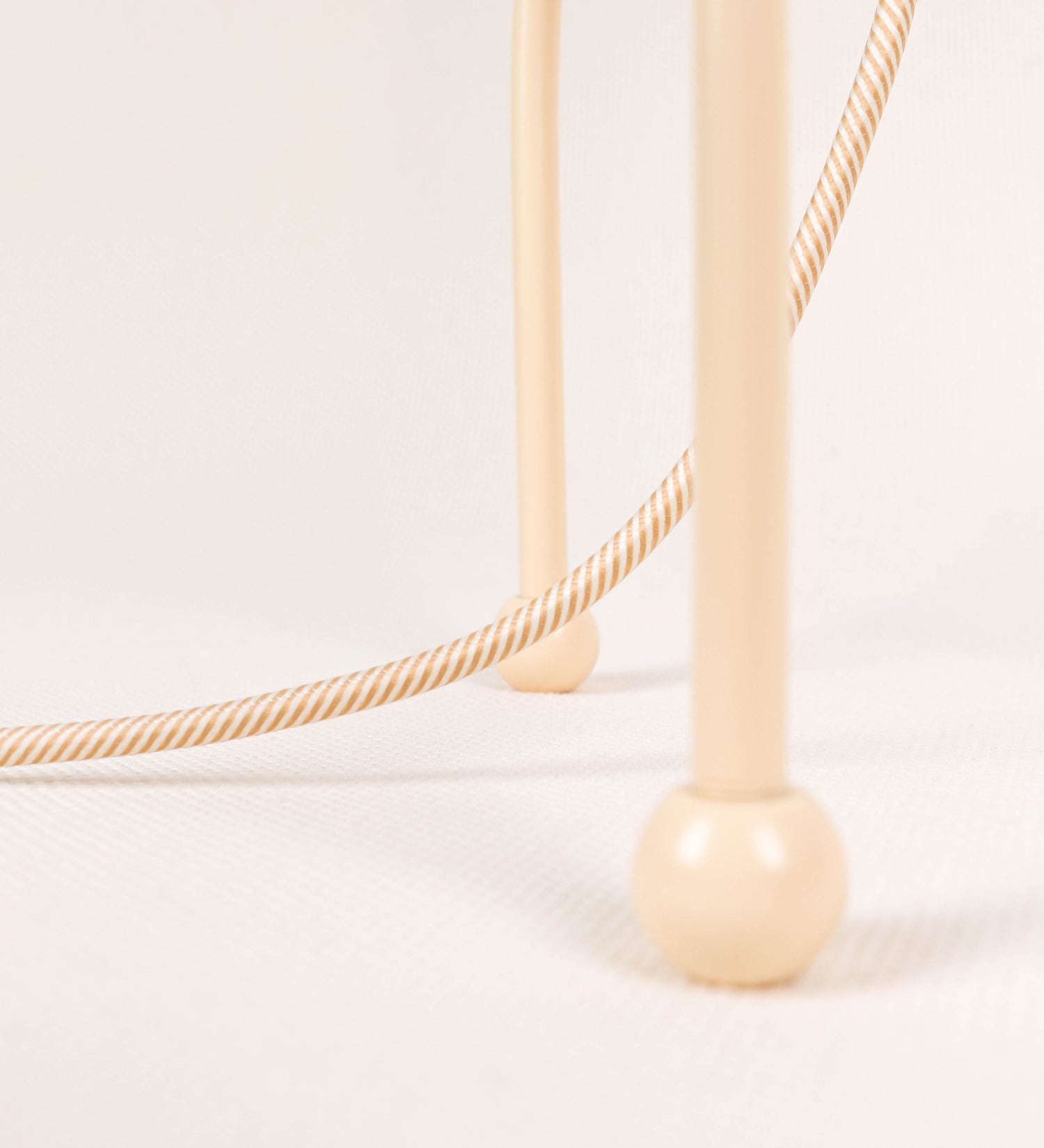 Candeeiro de mesa, de tom bege claro, com um design moderno e minimalista. O candeeiro tem o corpo com três pés curvados, terminando em pequenas esferas. A estrutura dos pés é interligada por uma peça central esférica. O cabo é revestido têxtil com um padrão de linhas douradas e brancas entrelaçadas, com interruptor e ficha transparentes.