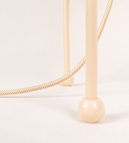 Candeeiro de mesa, de tom bege claro, com um design moderno e minimalista. O candeeiro tem o corpo com três pés curvados, terminando em pequenas esferas. A estrutura dos pés é interligada por uma peça central esférica. O cabo é revestido têxtil com um padrão de linhas douradas e brancas entrelaçadas, com interruptor e ficha transparentes.