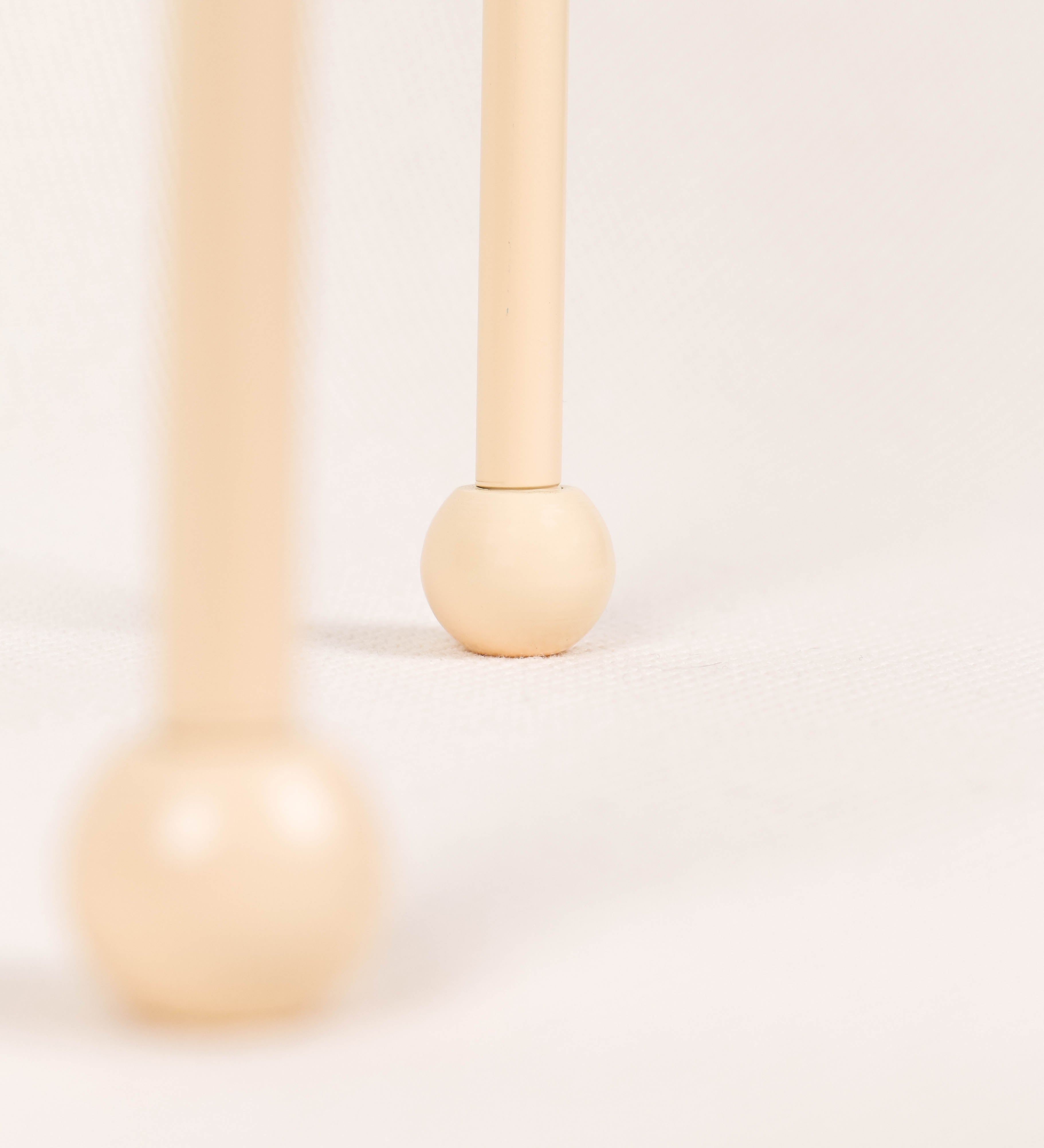 Candeeiro de mesa, de tom bege claro, com um design moderno e minimalista. O candeeiro tem o corpo com três pés curvados, terminando em pequenas esferas. A estrutura dos pés é interligada por uma peça central esférica. O cabo é revestido têxtil com um padrão de linhas douradas e brancas entrelaçadas, com interruptor e ficha transparentes.