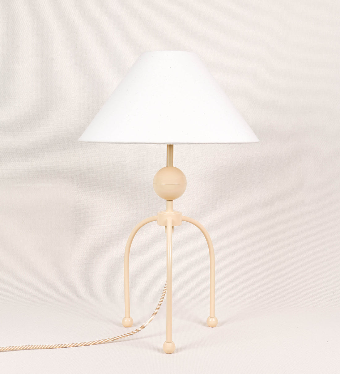 Candeeiro de mesa, de tom bege claro, com um design moderno e minimalista. O candeeiro tem o corpo com três pés curvados, terminando em pequenas esferas. A estrutura dos pés é interligada por uma peça central esférica. O cabo é revestido têxtil com um padrão de linhas douradas e brancas entrelaçadas, com interruptor e ficha transparentes. O abatjour é de forma cónica, em algodão, de tom bege claro com pequenas pintas escuras, semelhante à baunilha.