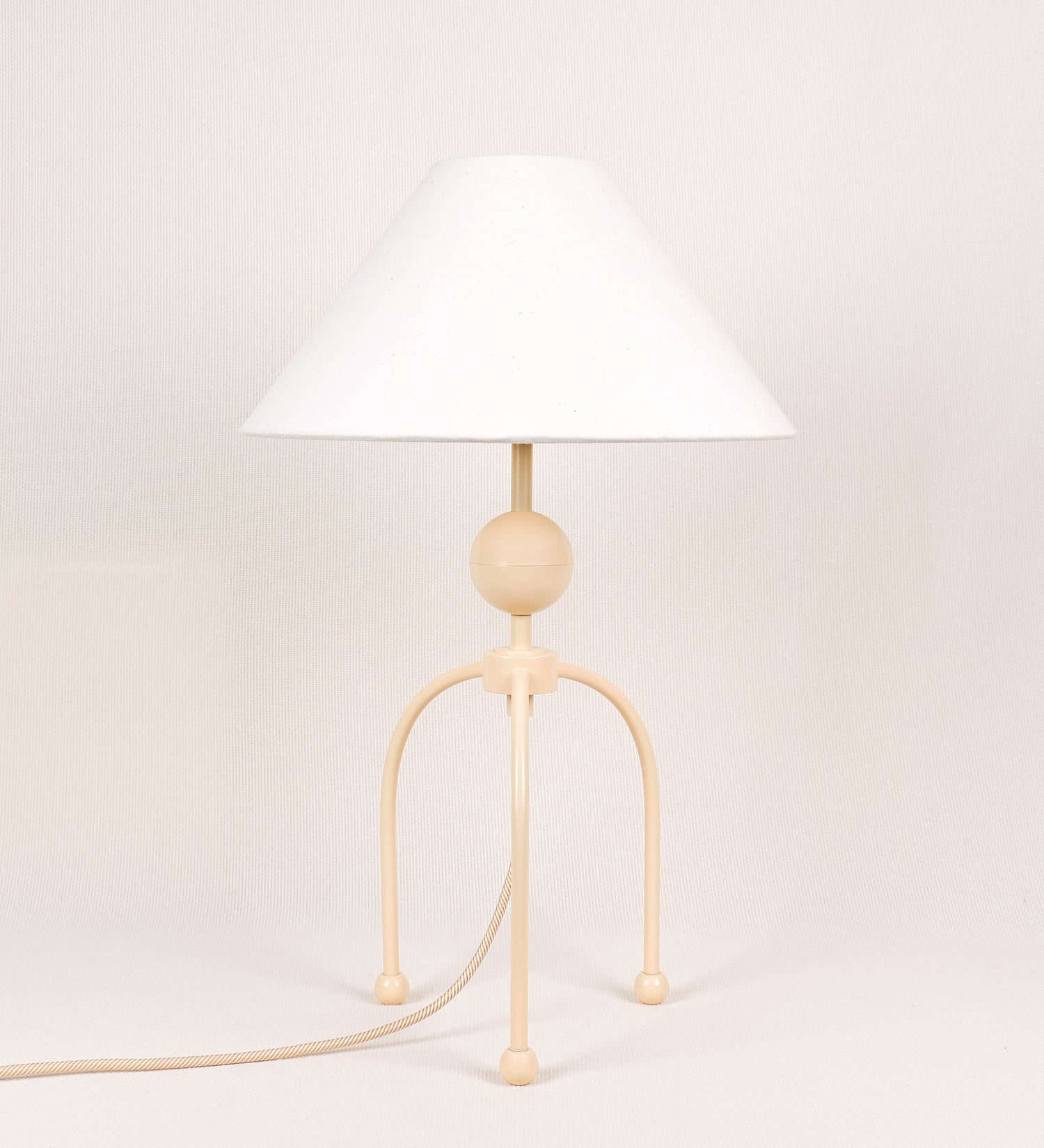 Candeeiro de mesa, de tom bege claro, com um design moderno e minimalista. O candeeiro tem o corpo com três pés curvados, terminando em pequenas esferas. A estrutura dos pés é interligada por uma peça central esférica. O cabo é revestido têxtil com um padrão de linhas douradas e brancas entrelaçadas, com interruptor e ficha transparentes. O abatjour é de forma cónica, em algodão, de tom bege claro com pequenas pintas escuras, semelhante à baunilha.