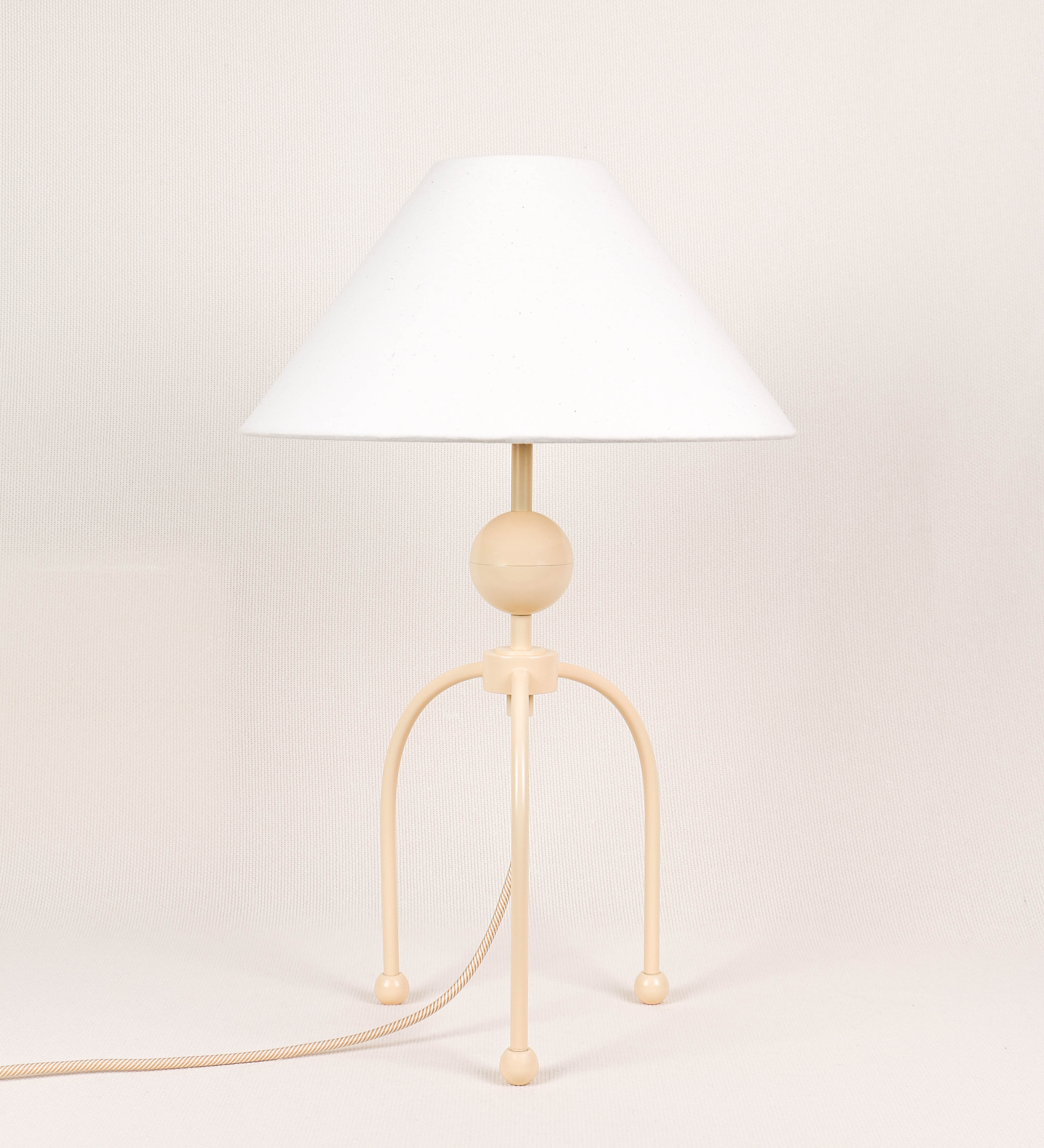 Candeeiro de mesa, de tom bege claro, com um design moderno e minimalista. O candeeiro tem o corpo com três pés curvados, terminando em pequenas esferas. A estrutura dos pés é interligada por uma peça central esférica. O cabo é revestido têxtil com um padrão de linhas douradas e brancas entrelaçadas, com interruptor e ficha transparentes. O abatjour é de forma cónica, em algodão, de tom bege claro com pequenas pintas escuras, semelhante à baunilha.