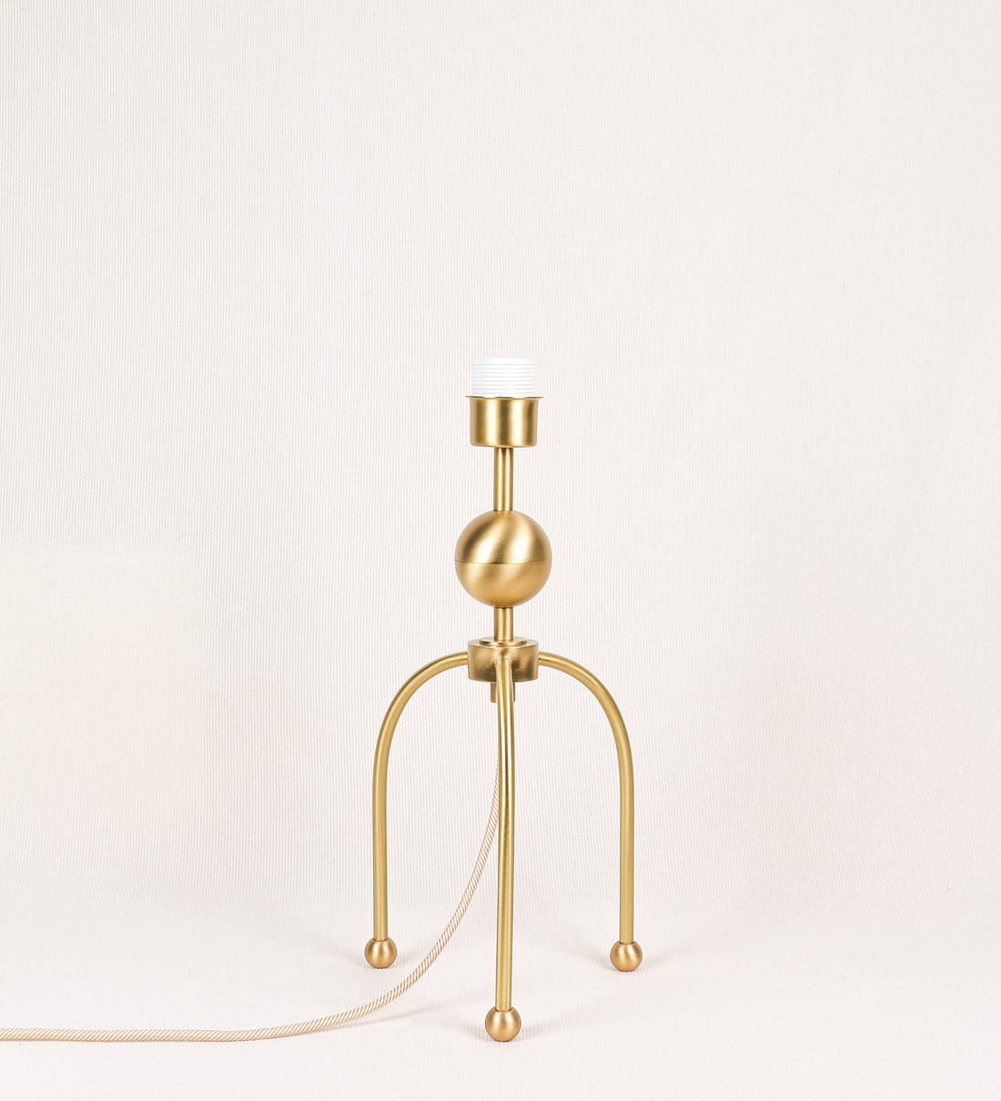 Candeeiro de mesa, em latão escovado, conferindo-lhe um tom dourado mate, com um design moderno e minimalista. O candeeiro tem o corpo com três pés curvados, terminando em pequenas esferas. A estrutura dos pés é interligada por uma peça central esférica. O cabo é revestido têxtil com um padrão de linhas douradas e brancas entrelaçadas, com interruptor e ficha transparentes.