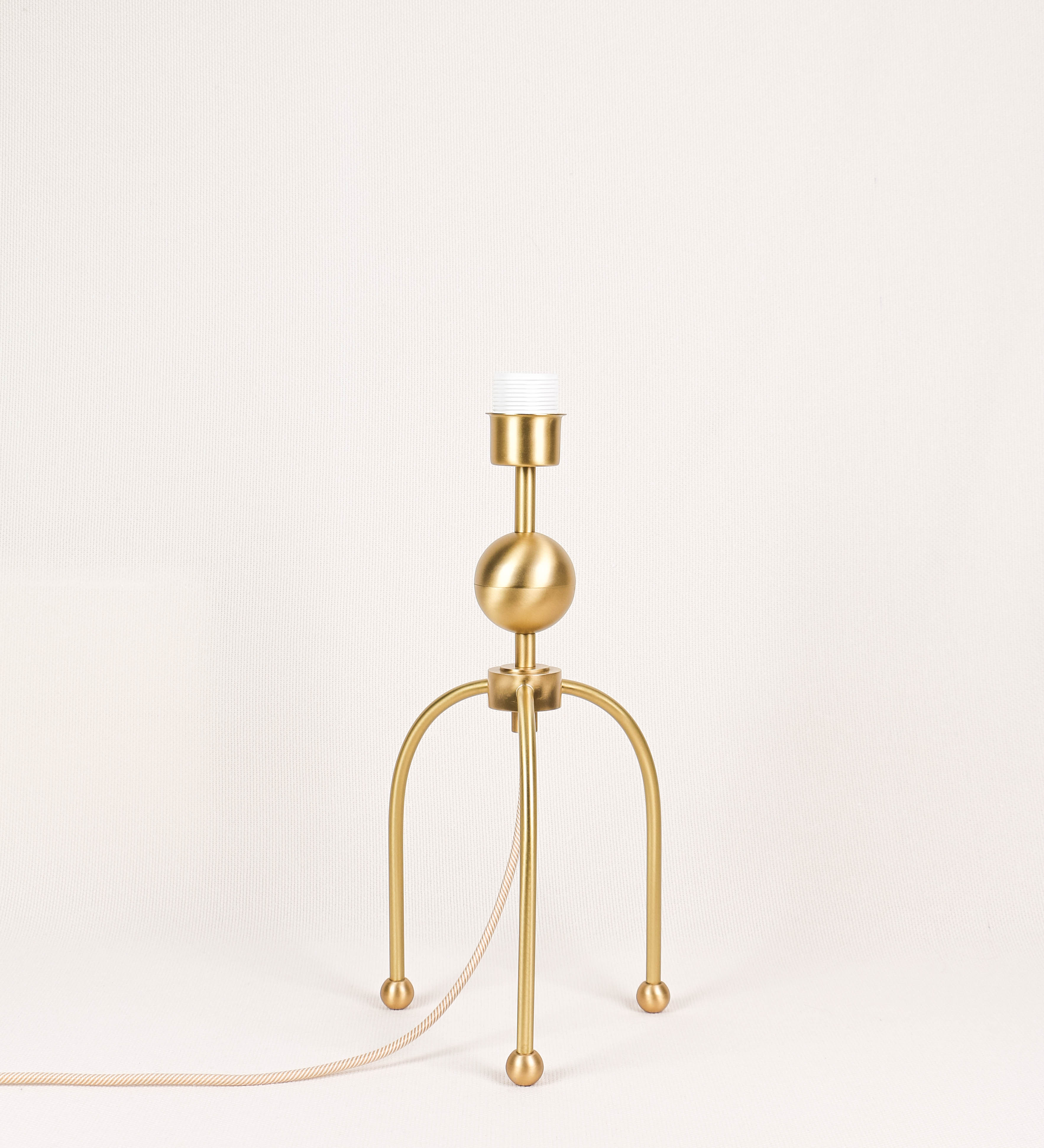 Candeeiro de mesa, em latão escovado, conferindo-lhe um tom dourado mate, com um design moderno e minimalista. O candeeiro tem o corpo com três pés curvados, terminando em pequenas esferas. A estrutura dos pés é interligada por uma peça central esférica. O cabo é revestido têxtil com um padrão de linhas douradas e brancas entrelaçadas, com interruptor e ficha transparentes.
