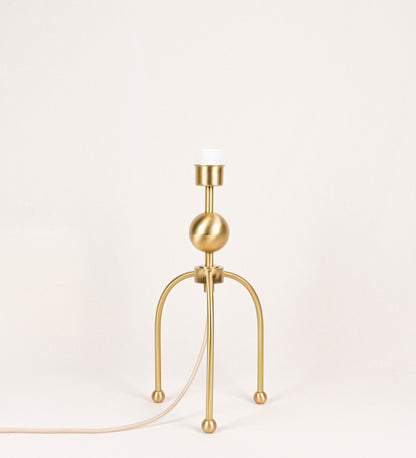 Candeeiro de mesa, em latão escovado, conferindo-lhe um tom dourado mate, com um design moderno e minimalista. O candeeiro tem o corpo com três pés curvados, terminando em pequenas esferas. A estrutura dos pés é interligada por uma peça central esférica. O cabo é revestido têxtil com um padrão de linhas douradas e brancas entrelaçadas, com interruptor e ficha transparentes.