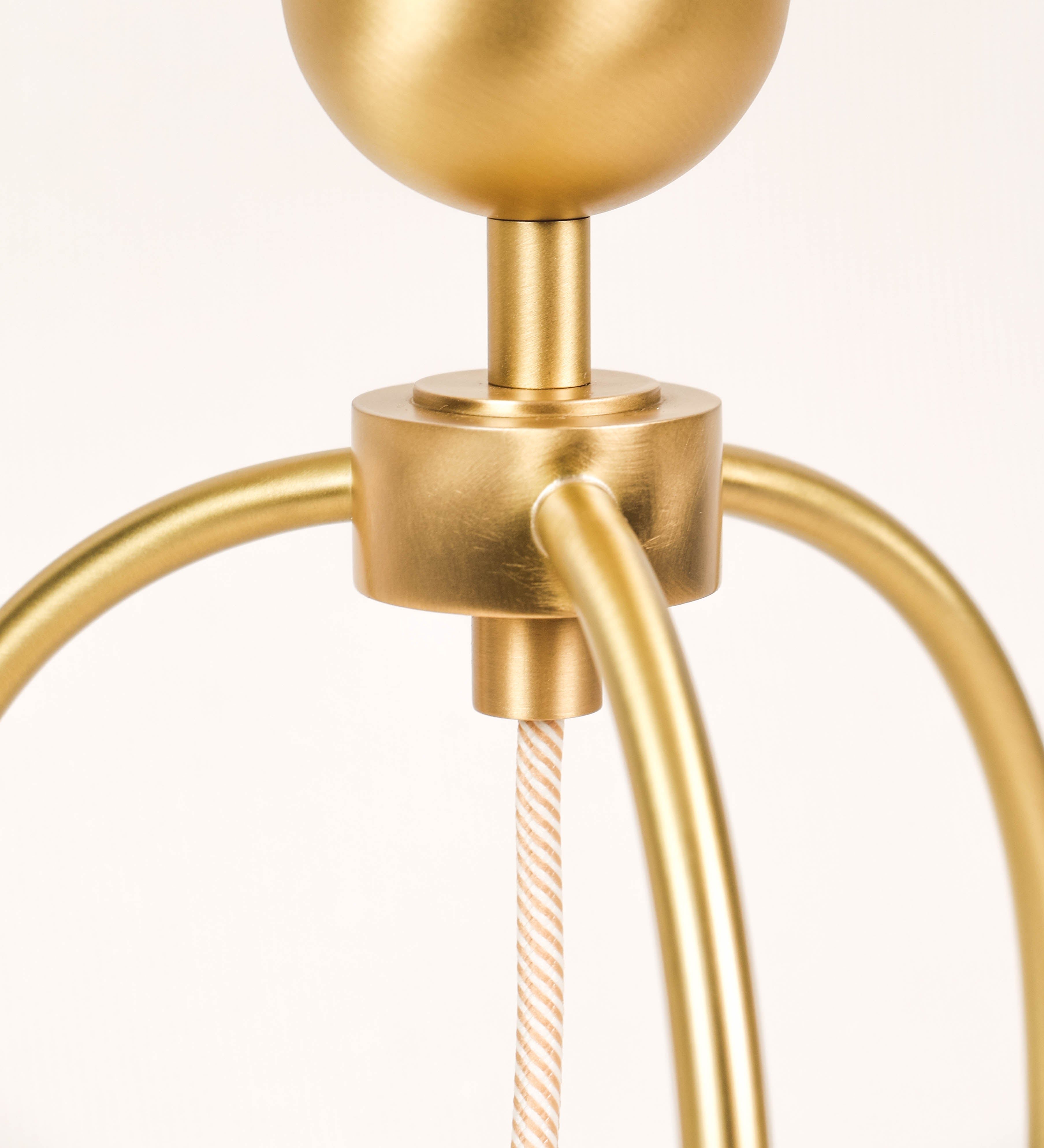 Candeeiro de mesa, em latão escovado, conferindo-lhe um tom dourado mate, com um design moderno e minimalista. O candeeiro tem o corpo com três pés curvados, terminando em pequenas esferas. A estrutura dos pés é interligada por uma peça central esférica. O cabo é revestido têxtil com um padrão de linhas douradas e brancas entrelaçadas, com interruptor e ficha transparentes. 