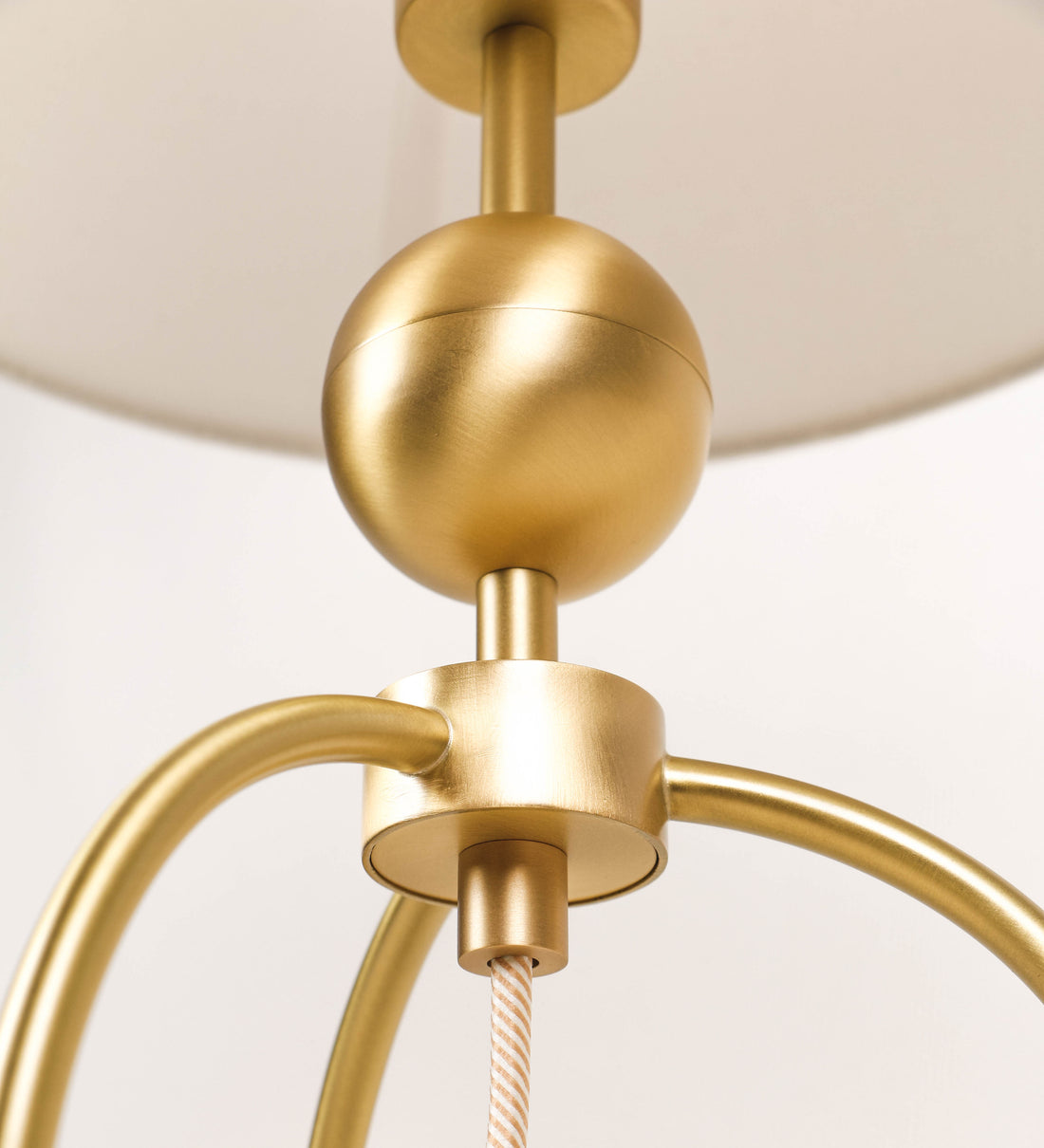 Candeeiro de mesa, em latão escovado, conferindo-lhe um tom dourado mate, com um design moderno e minimalista. O candeeiro tem o corpo com três pés curvados, terminando em pequenas esferas. A estrutura dos pés é interligada por uma peça central esférica. O cabo é revestido têxtil com um padrão de linhas douradas e brancas entrelaçadas, com interruptor e ficha transparentes. O abatjour é de forma cónica, em algodão, de tom bege claro com pequenas pintas escuras, semelhante à baunilha.