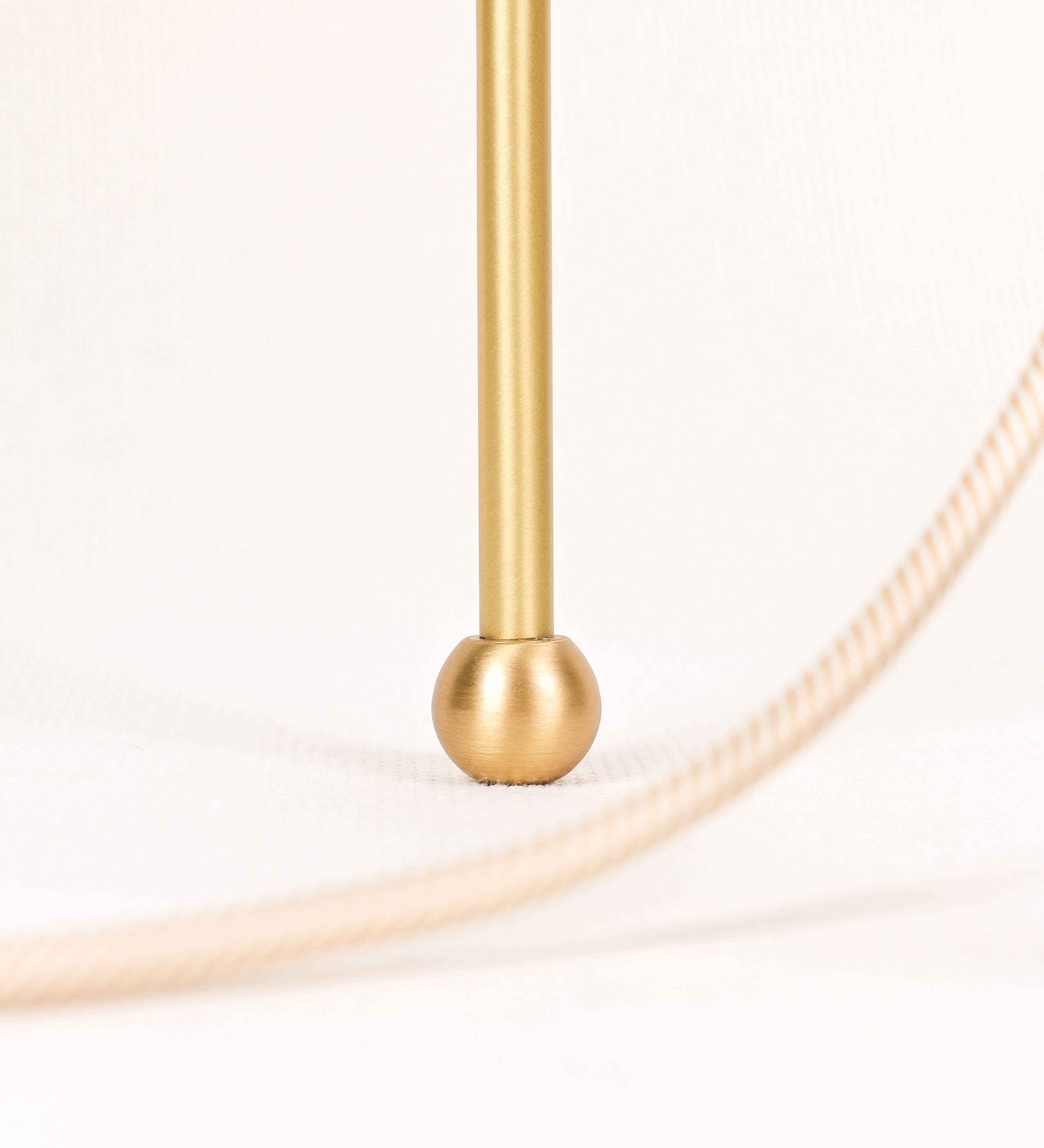 Candeeiro de mesa, em latão escovado, conferindo-lhe um tom dourado mate, com um design moderno e minimalista. O candeeiro tem o corpo com três pés curvados, terminando em pequenas esferas. A estrutura dos pés é interligada por uma peça central esférica. O cabo é revestido têxtil com um padrão de linhas douradas e brancas entrelaçadas, com interruptor e ficha transparentes. 