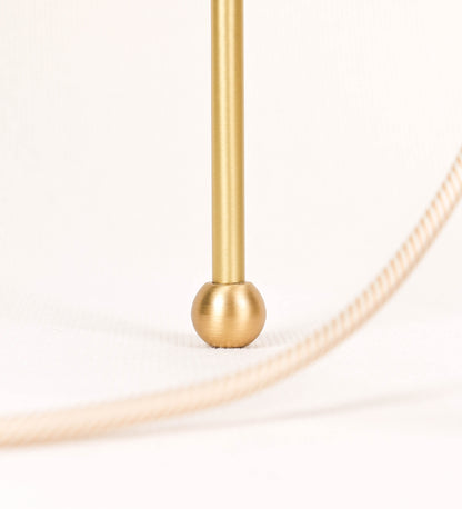 Candeeiro de mesa, em latão escovado, conferindo-lhe um tom dourado mate, com um design moderno e minimalista. O candeeiro tem o corpo com três pés curvados, terminando em pequenas esferas. A estrutura dos pés é interligada por uma peça central esférica. O cabo é revestido têxtil com um padrão de linhas douradas e brancas entrelaçadas, com interruptor e ficha transparentes. 