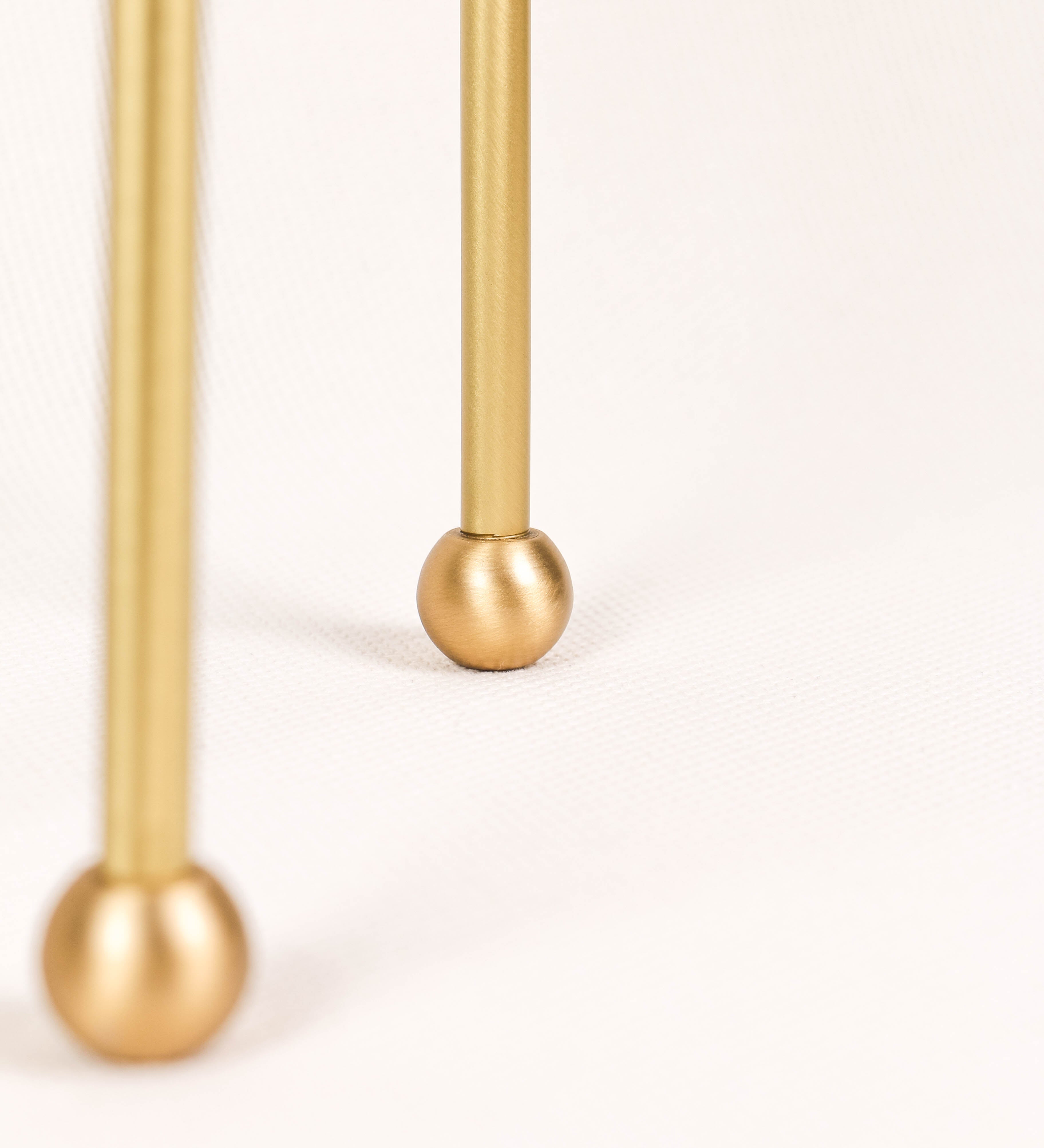 Candeeiro de mesa, em latão escovado, conferindo-lhe um tom dourado mate, com um design moderno e minimalista. O candeeiro tem o corpo com três pés curvados, terminando em pequenas esferas. A estrutura dos pés é interligada por uma peça central esférica. O cabo é revestido têxtil com um padrão de linhas douradas e brancas entrelaçadas, com interruptor e ficha transparentes.