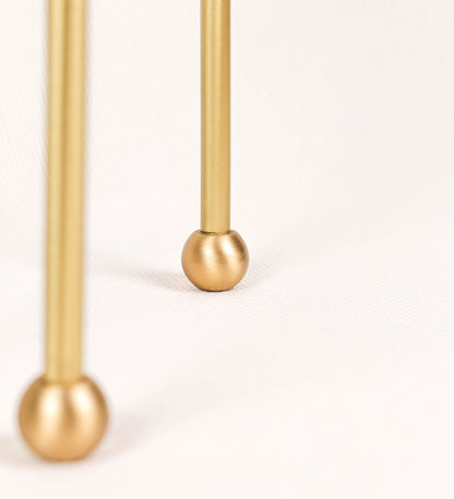 Candeeiro de mesa, em latão escovado, conferindo-lhe um tom dourado mate, com um design moderno e minimalista. O candeeiro tem o corpo com três pés curvados, terminando em pequenas esferas. A estrutura dos pés é interligada por uma peça central esférica. O cabo é revestido têxtil com um padrão de linhas douradas e brancas entrelaçadas, com interruptor e ficha transparentes.