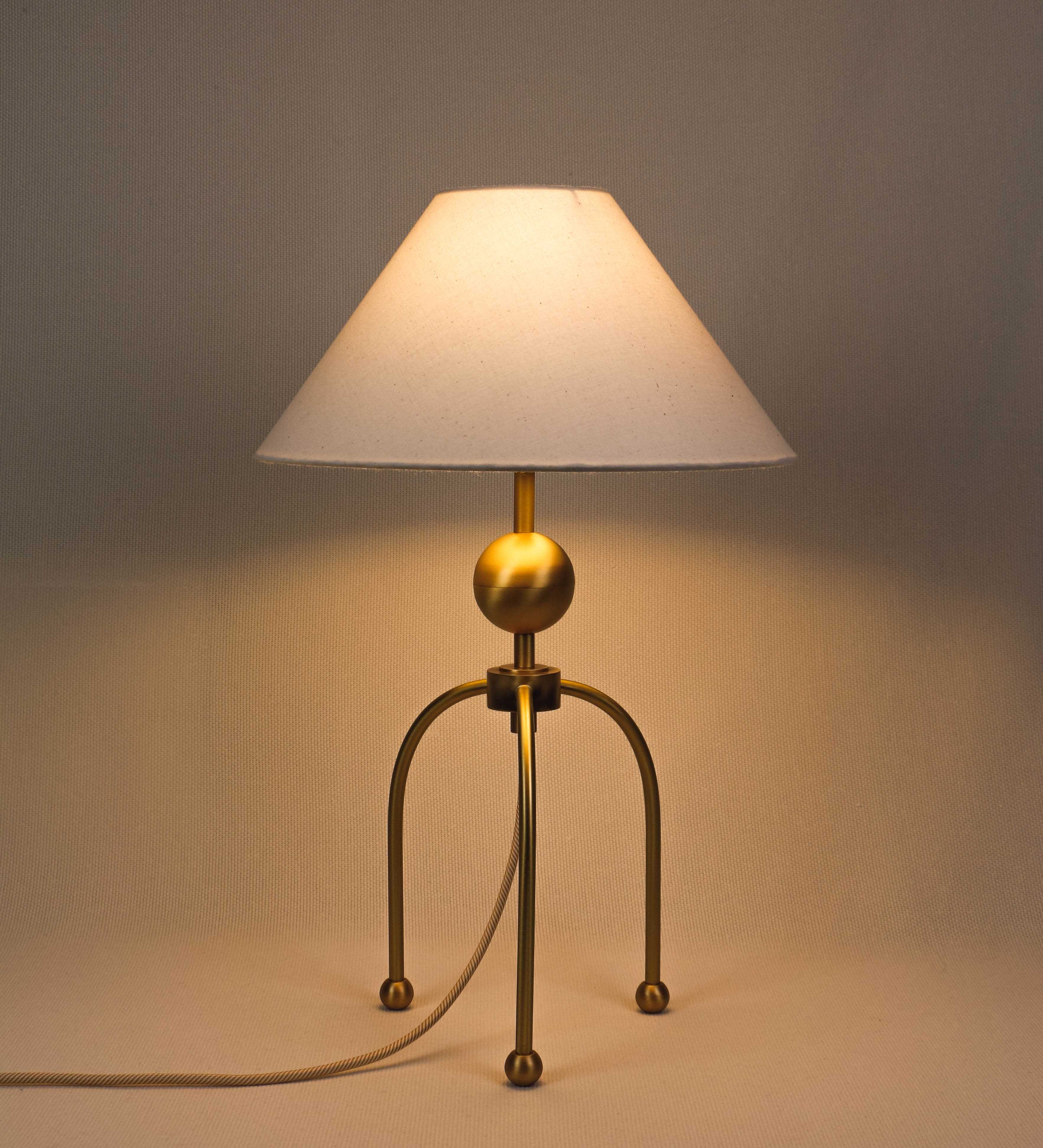 Candeeiro de mesa, em latão escovado, conferindo-lhe um tom dourado mate, com um design moderno e minimalista. O candeeiro tem o corpo com três pés curvados, terminando em pequenas esferas. A estrutura dos pés é interligada por uma peça central esférica. O cabo é revestido têxtil com um padrão de linhas douradas e brancas entrelaçadas, com interruptor e ficha transparentes. O abatjour é de forma cónica, em algodão, de tom bege claro com pequenas pintas escuras, semelhante à baunilha.