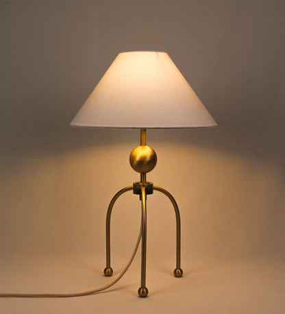 Candeeiro de mesa, em latão escovado, conferindo-lhe um tom dourado mate, com um design moderno e minimalista. O candeeiro tem o corpo com três pés curvados, terminando em pequenas esferas. A estrutura dos pés é interligada por uma peça central esférica. O cabo é revestido têxtil com um padrão de linhas douradas e brancas entrelaçadas, com interruptor e ficha transparentes. O abatjour é de forma cónica, em algodão, de tom bege claro com pequenas pintas escuras, semelhante à baunilha.