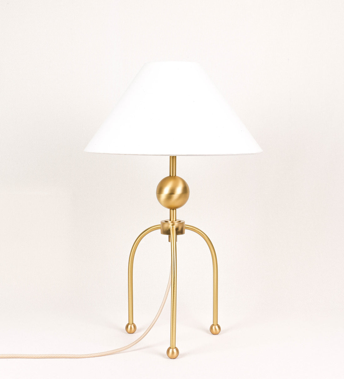 Candeeiro de mesa, em latão escovado, conferindo-lhe um tom dourado mate, com um design moderno e minimalista. O candeeiro tem o corpo com três pés curvados, terminando em pequenas esferas. A estrutura dos pés é interligada por uma peça central esférica. O cabo é revestido têxtil com um padrão de linhas douradas e brancas entrelaçadas, com interruptor e ficha transparentes. O abatjour é de forma cónica, em algodão, de tom bege claro com pequenas pintas escuras, semelhante à baunilha.