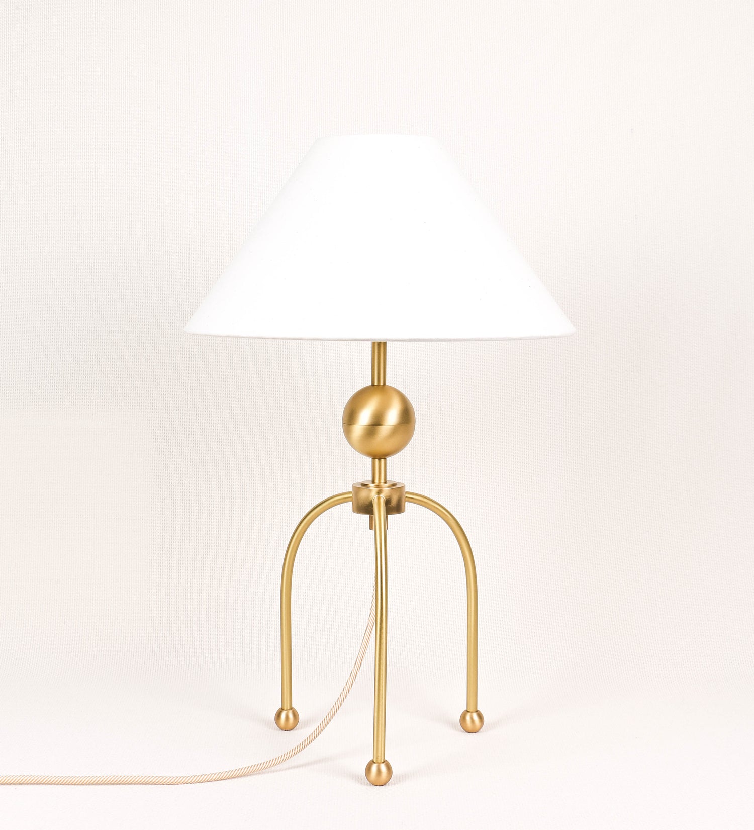Candeeiro de mesa, em latão escovado, conferindo-lhe um tom dourado mate, com um design moderno e minimalista. O candeeiro tem o corpo com três pés curvados, terminando em pequenas esferas. A estrutura dos pés é interligada por uma peça central esférica. O cabo é revestido têxtil com um padrão de linhas douradas e brancas entrelaçadas, com interruptor e ficha transparentes. O abatjour é de forma cónica, em algodão, de tom bege claro com pequenas pintas escuras, semelhante à baunilha.