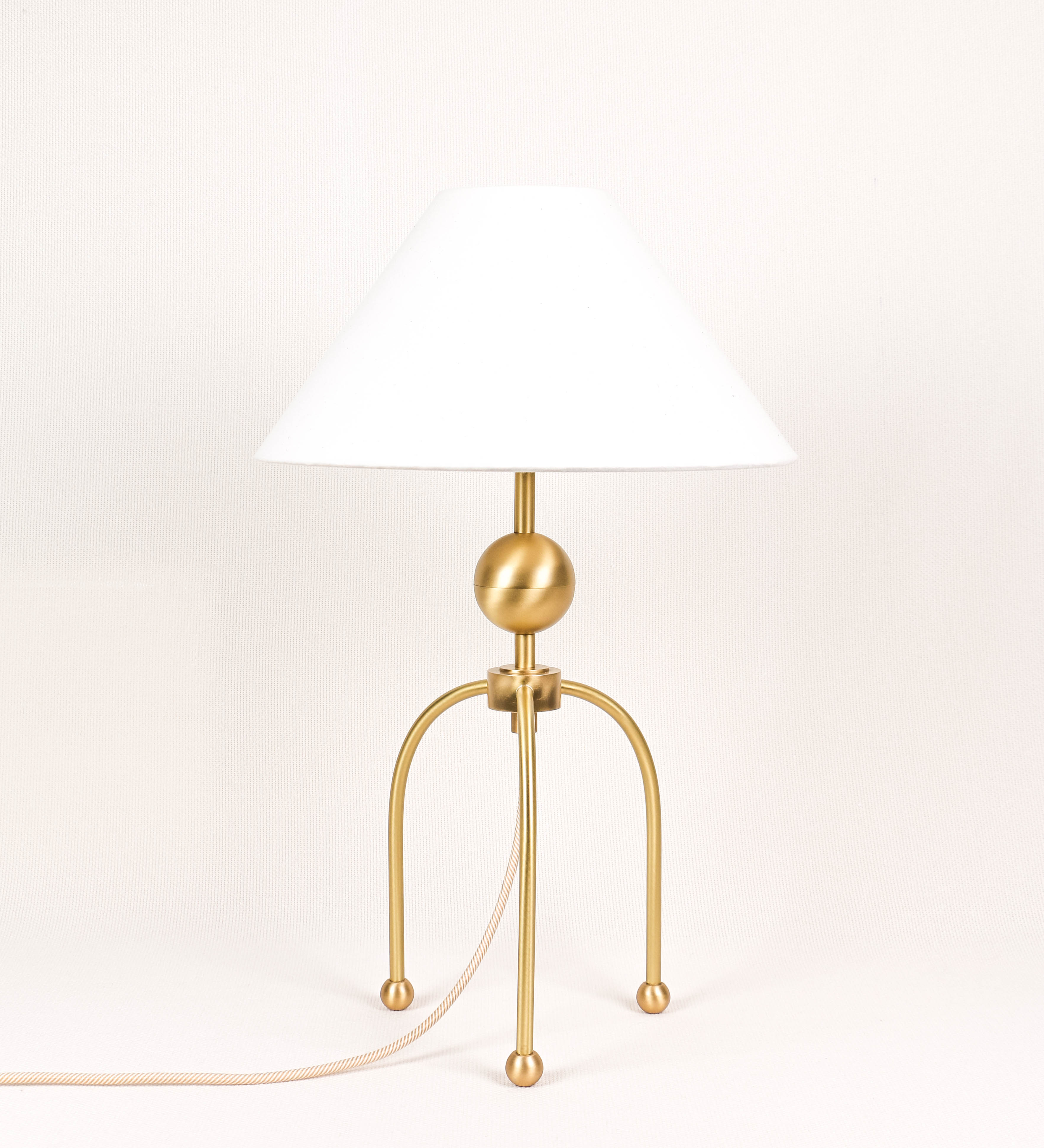 Candeeiro de mesa, em latão escovado, conferindo-lhe um tom dourado mate, com um design moderno e minimalista. O candeeiro tem o corpo com três pés curvados, terminando em pequenas esferas. A estrutura dos pés é interligada por uma peça central esférica. O cabo é revestido têxtil com um padrão de linhas douradas e brancas entrelaçadas, com interruptor e ficha transparentes. O abatjour é de forma cónica, em algodão, de tom bege claro com pequenas pintas escuras, semelhante à baunilha.