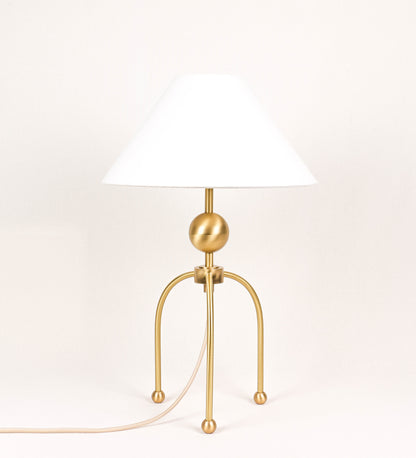 Candeeiro de mesa, em latão escovado, conferindo-lhe um tom dourado mate, com um design moderno e minimalista. O candeeiro tem o corpo com três pés curvados, terminando em pequenas esferas. A estrutura dos pés é interligada por uma peça central esférica. O cabo é revestido têxtil com um padrão de linhas douradas e brancas entrelaçadas, com interruptor e ficha transparentes. O abatjour é de forma cónica, em algodão, de tom bege claro com pequenas pintas escuras, semelhante à baunilha.