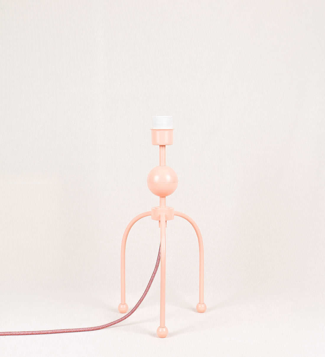 Candeeiro de mesa, de tom rosa salmão, com um design moderno e minimalista. O candeeiro tem o corpo com três pés curvados, terminando em pequenas esferas. A estrutura dos pés é interligada por uma peça central esférica. O cabo é revestido têxtil de tom rosa velho, com interruptor e ficha transparentes.