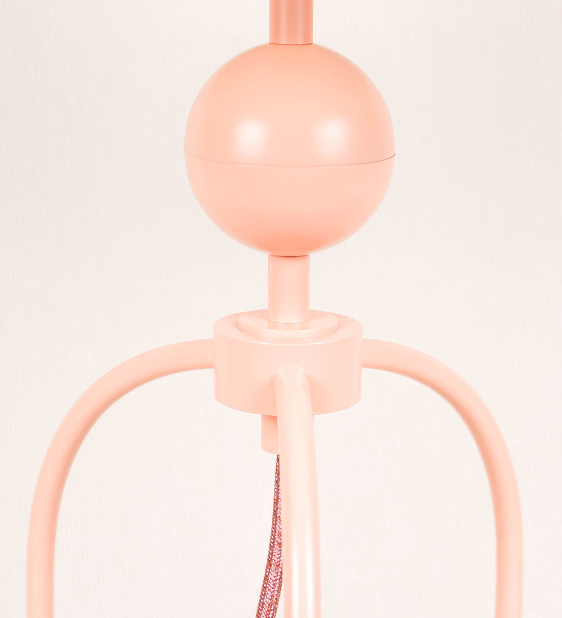 Candeeiro de mesa, de tom rosa salmão, com um design moderno e minimalista. O candeeiro tem o corpo com três pés curvados, terminando em pequenas esferas. A estrutura dos pés é interligada por uma peça central esférica. O cabo é revestido têxtil de tom rosa velho, com interruptor e ficha transparentes.