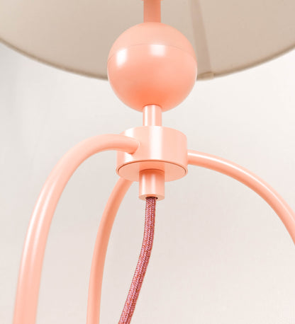 Candeeiro de mesa, de tom rosa salmão, com um design moderno e minimalista. O candeeiro tem o corpo com três pés curvados, terminando em pequenas esferas. A estrutura dos pés é interligada por uma peça central esférica. O cabo é revestido têxtil de tom rosa velho, com interruptor e ficha transparentes. O abatjour é de forma cónica, em algodão, de tom bege claro com pequenas pintas escuras, semelhante à baunilha.
