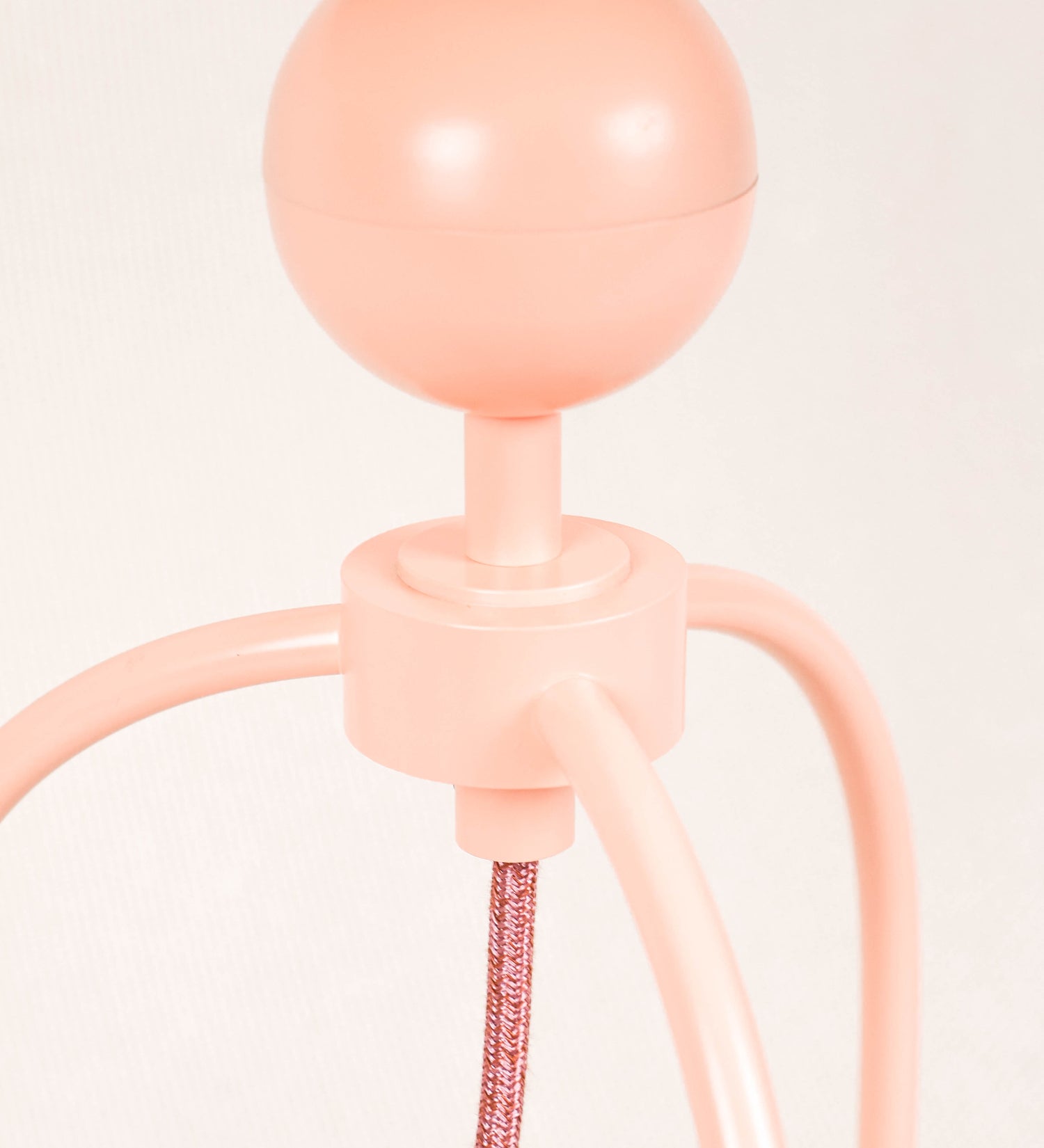 Candeeiro de mesa, de tom rosa salmão, com um design moderno e minimalista. O candeeiro tem o corpo com três pés curvados, terminando em pequenas esferas. A estrutura dos pés é interligada por uma peça central esférica. O cabo é revestido têxtil de tom rosa velho, com interruptor e ficha transparentes. O abatjour é de forma cónica, em algodão, de tom bege claro com pequenas pintas escuras, semelhante à baunilha.