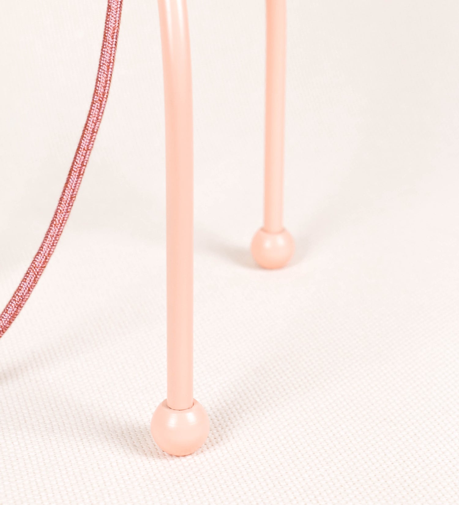 Candeeiro de mesa, de tom rosa salmão, com um design moderno e minimalista. O candeeiro tem o corpo com três pés curvados, terminando em pequenas esferas. A estrutura dos pés é interligada por uma peça central esférica. O cabo é revestido têxtil de tom rosa velho, com interruptor e ficha transparentes. O abatjour é de forma cónica, em algodão, de tom bege claro com pequenas pintas escuras, semelhante à baunilha.