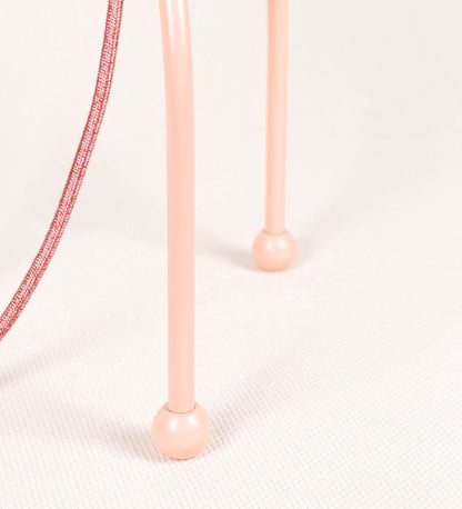 Candeeiro de mesa, de tom rosa salmão, com um design moderno e minimalista. O candeeiro tem o corpo com três pés curvados, terminando em pequenas esferas. A estrutura dos pés é interligada por uma peça central esférica. O cabo é revestido têxtil de tom rosa velho, com interruptor e ficha transparentes. O abatjour é de forma cónica, em algodão, de tom bege claro com pequenas pintas escuras, semelhante à baunilha.