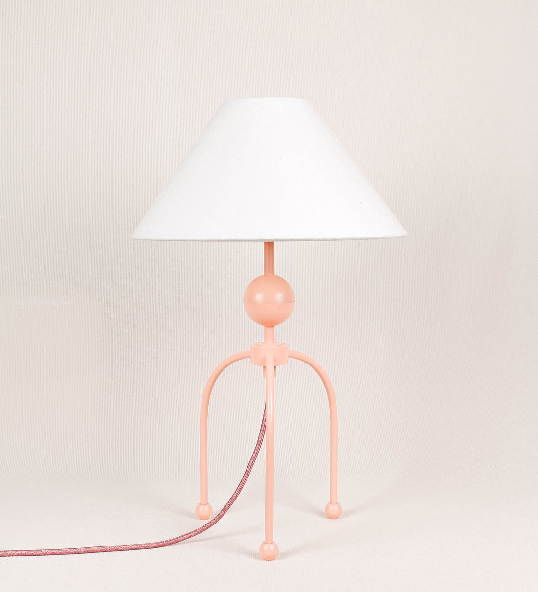 Candeeiro de mesa, de tom rosa salmão, com um design moderno e minimalista. O candeeiro tem o corpo com três pés curvados, terminando em pequenas esferas. A estrutura dos pés é interligada por uma peça central esférica. O cabo é revestido têxtil de tom rosa velho, com interruptor e ficha transparentes. O abatjour é de forma cónica, em algodão, de tom bege claro com pequenas pintas escuras, semelhante à baunilha.