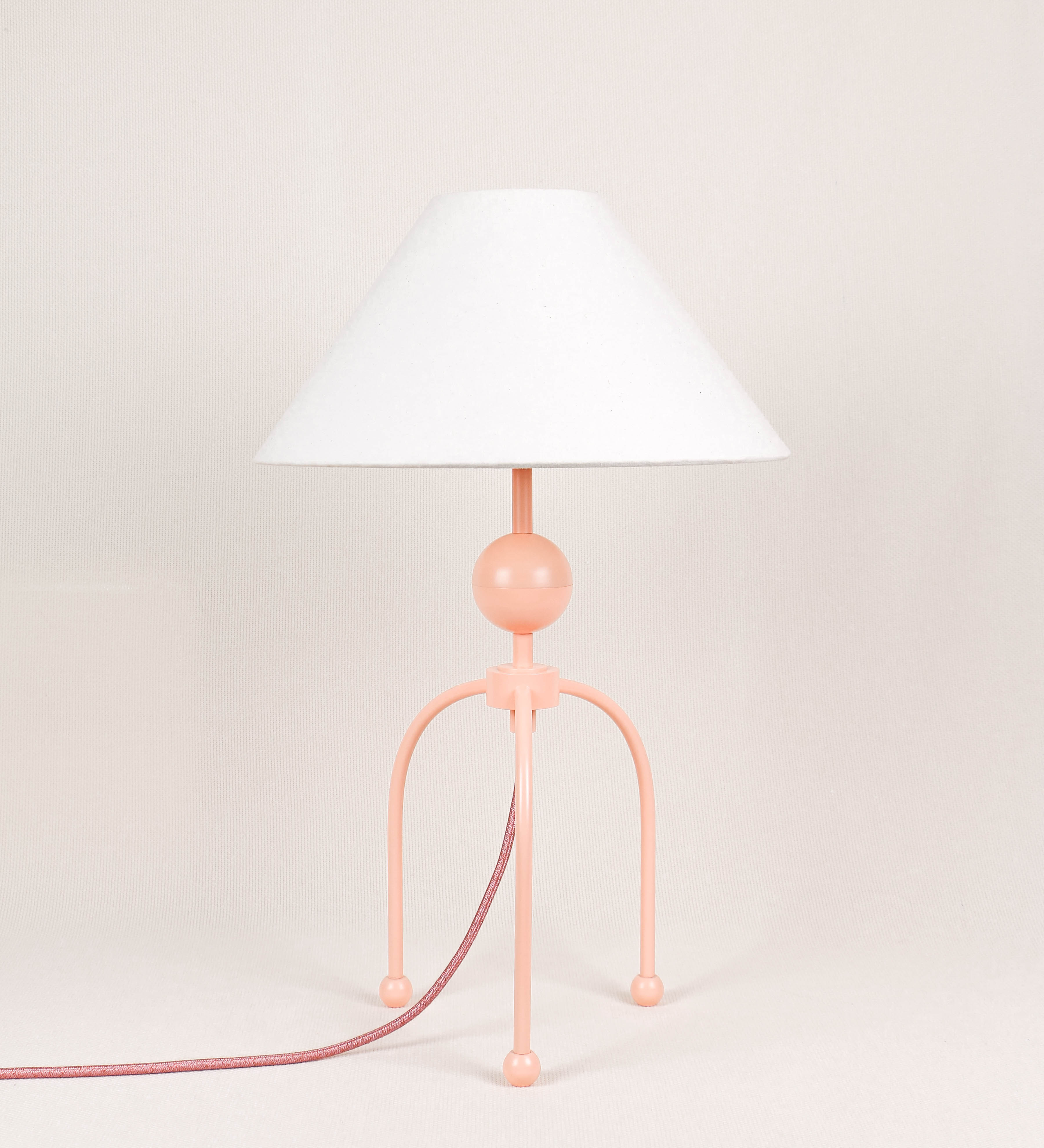 Candeeiro de mesa, de tom rosa salmão, com um design moderno e minimalista. O candeeiro tem o corpo com três pés curvados, terminando em pequenas esferas. A estrutura dos pés é interligada por uma peça central esférica. O cabo é revestido têxtil de tom rosa velho, com interruptor e ficha transparentes. O abatjour é de forma cónica, em algodão, de tom bege claro com pequenas pintas escuras, semelhante à baunilha.