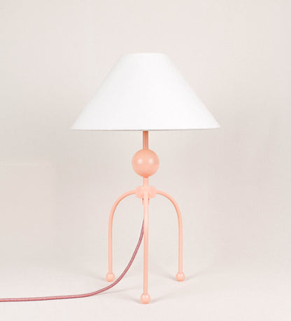 Candeeiro de mesa, de tom rosa salmão, com um design moderno e minimalista. O candeeiro tem o corpo com três pés curvados, terminando em pequenas esferas. A estrutura dos pés é interligada por uma peça central esférica. O cabo é revestido têxtil de tom rosa velho, com interruptor e ficha transparentes. O abatjour é de forma cónica, em algodão, de tom bege claro com pequenas pintas escuras, semelhante à baunilha.